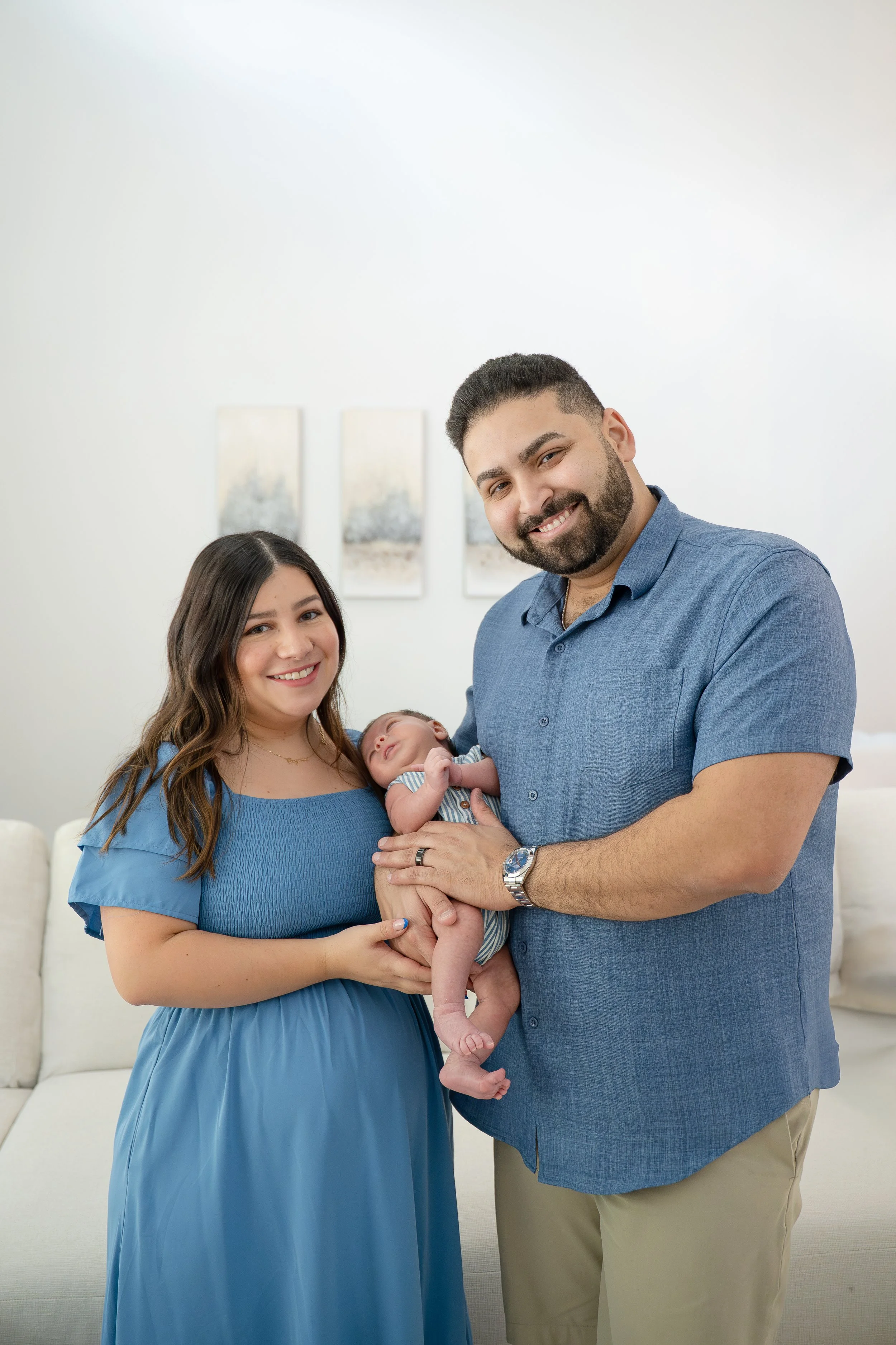 A happy family of four in a living room: woman in a blue dress, man in a blue shirt holding a baby, and girl smiling, in their The Woodlands home.