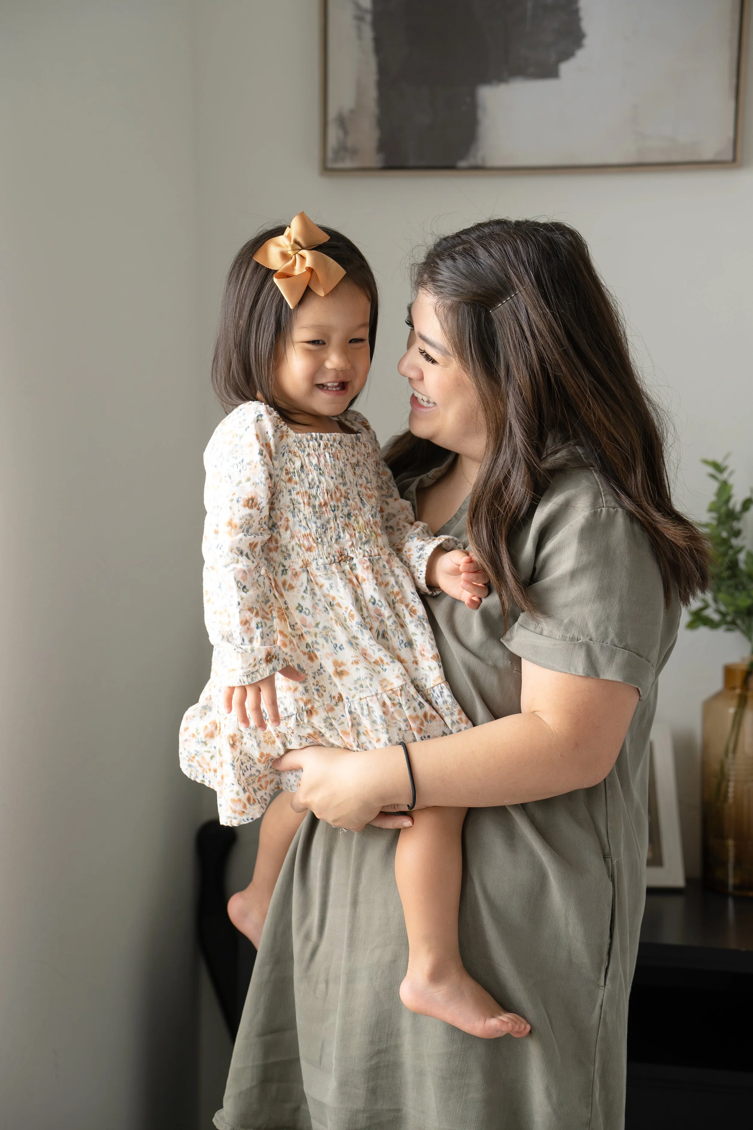 A woman with long dark hair, smiling, holds a young girl with a bow in her hair, wearing a floral dress, as they smile and look at each other at home in Katy, TX.