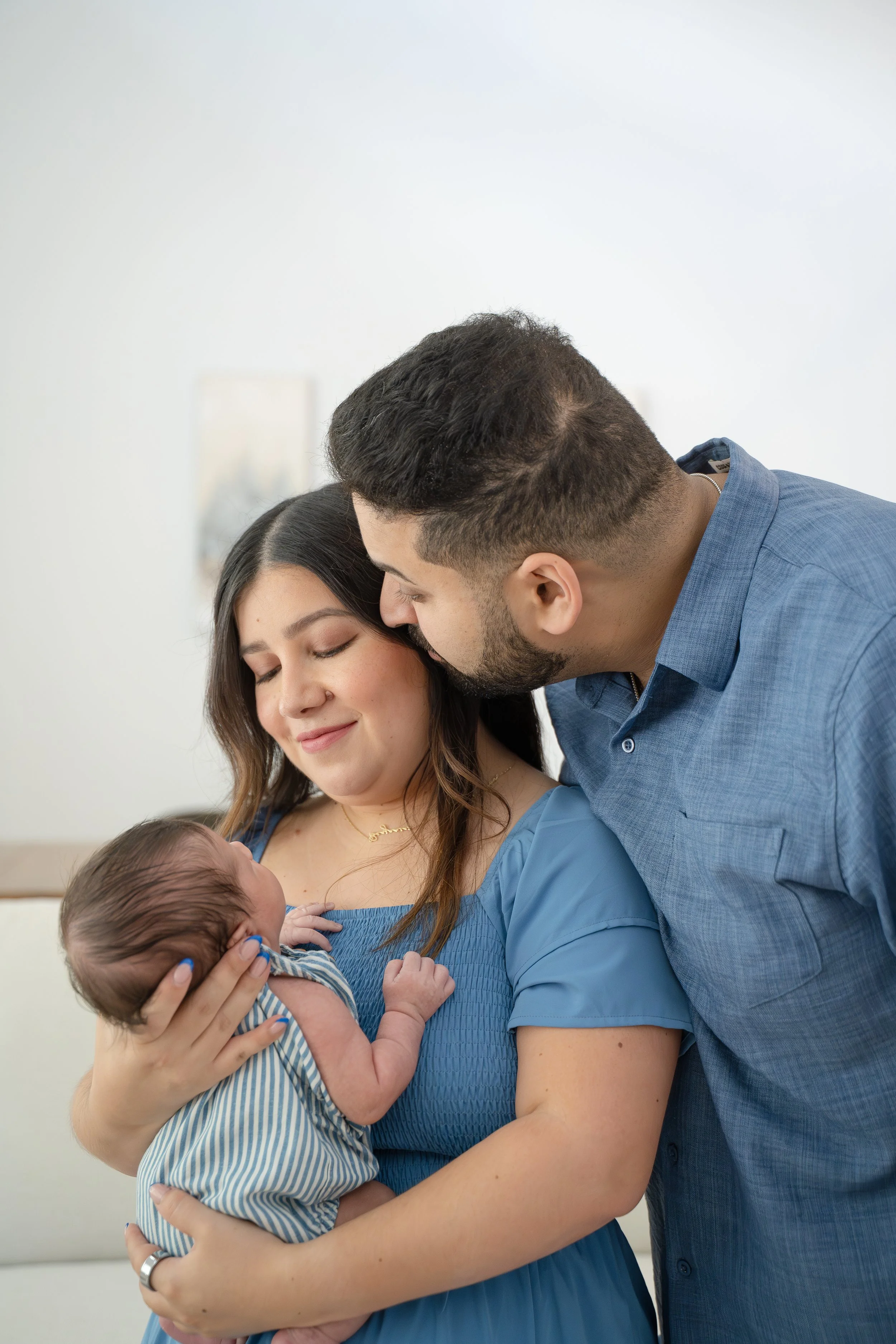 A young couple holding their newborn baby, with the mother smiling and the father leaning in closely, in a well-lit indoor setting, in their The Woodlands home.