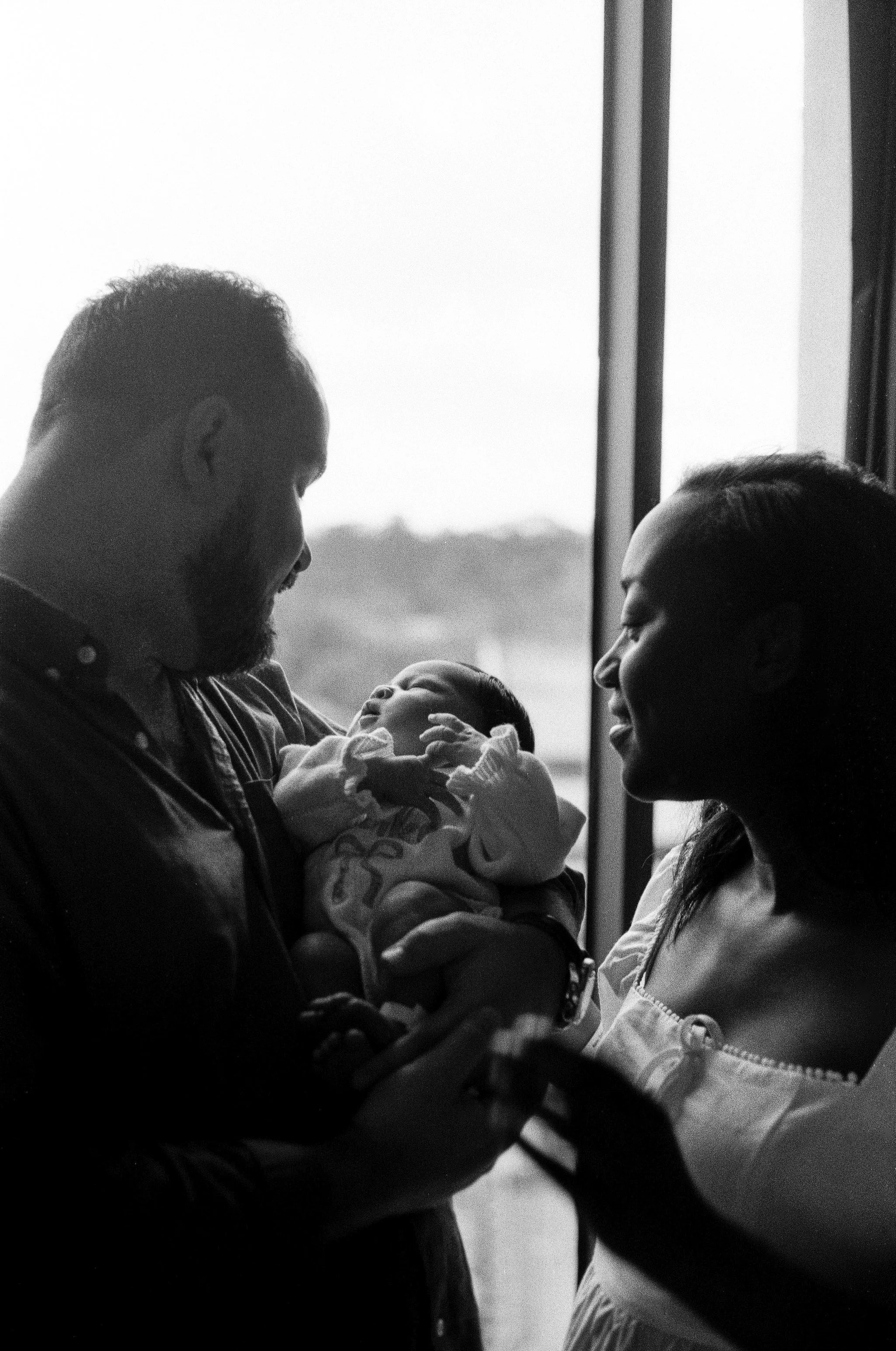 A black and white photo of a man and a woman looking at a sleeping baby. The man is on the left, holding the baby, and the woman is on the right, smiling at the baby. They are indoors near a window, in the Houston Ruby Postnatal. 