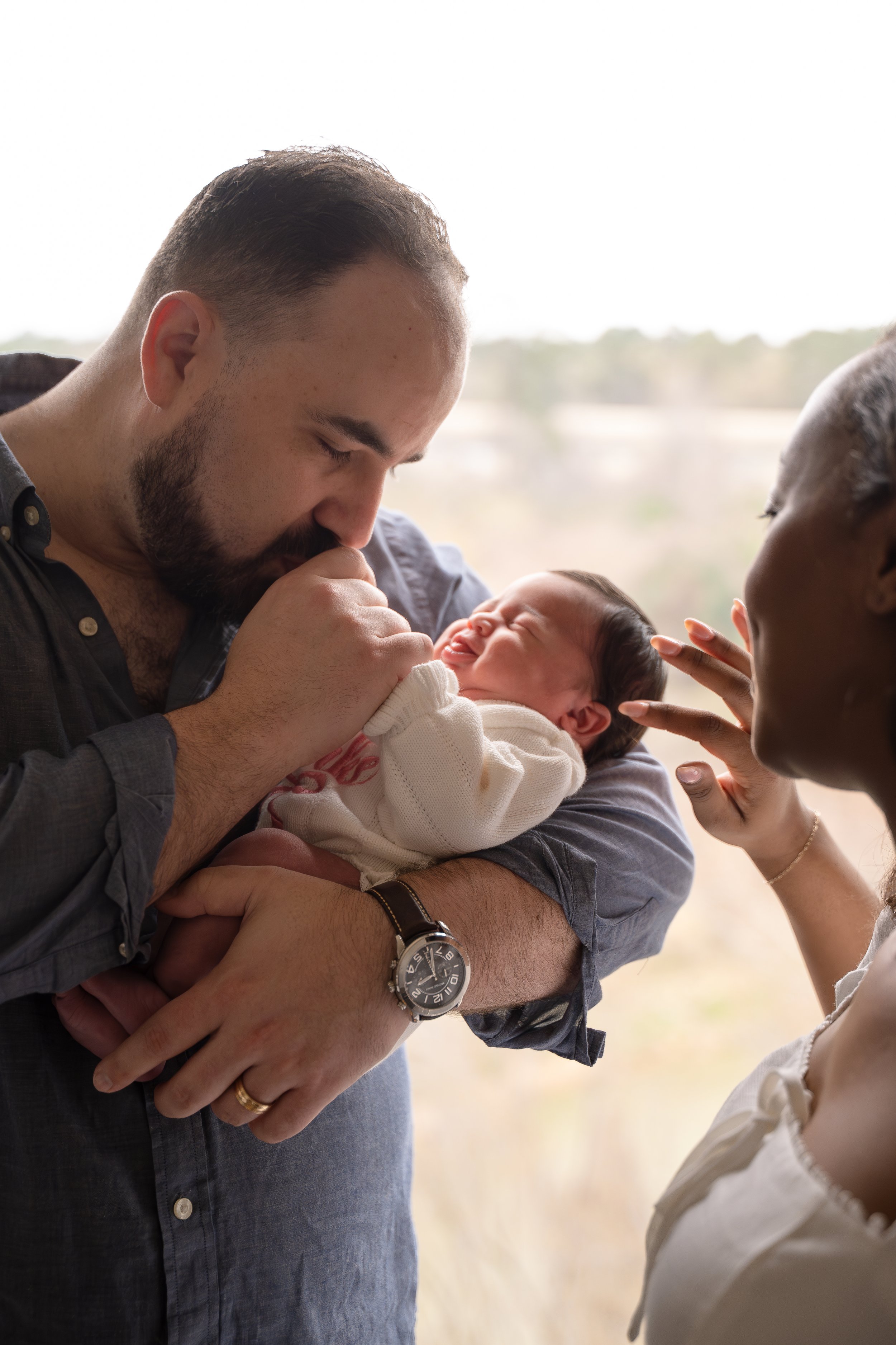 A man holding a baby with a woman standing nearby, in front of a window with a view of a landscape, in the Houston Ruby Postnatal. 