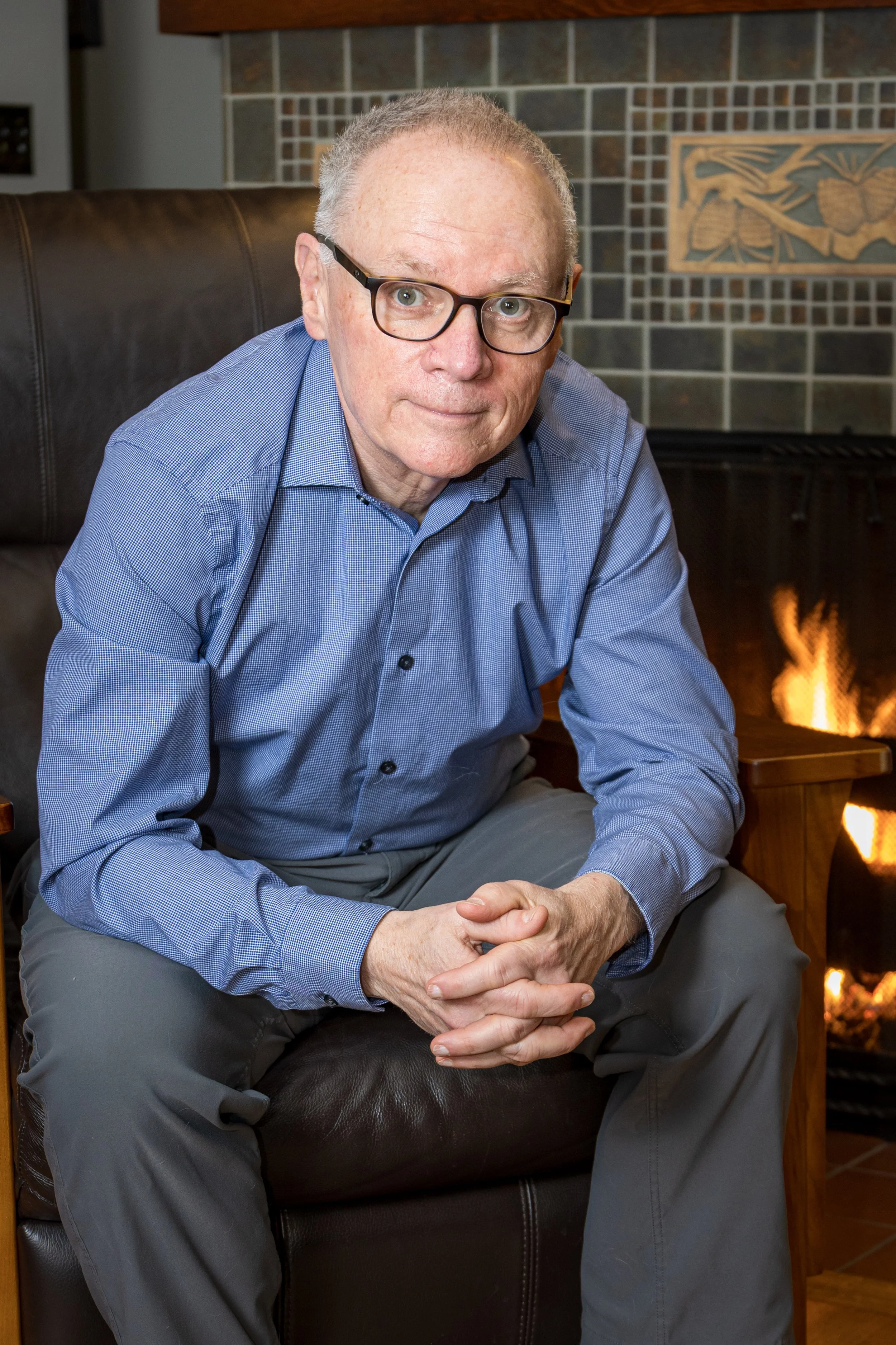 David Womack, wearing a blue dress shirt and gray pants, sitting on a leather chair near a fireplace.
