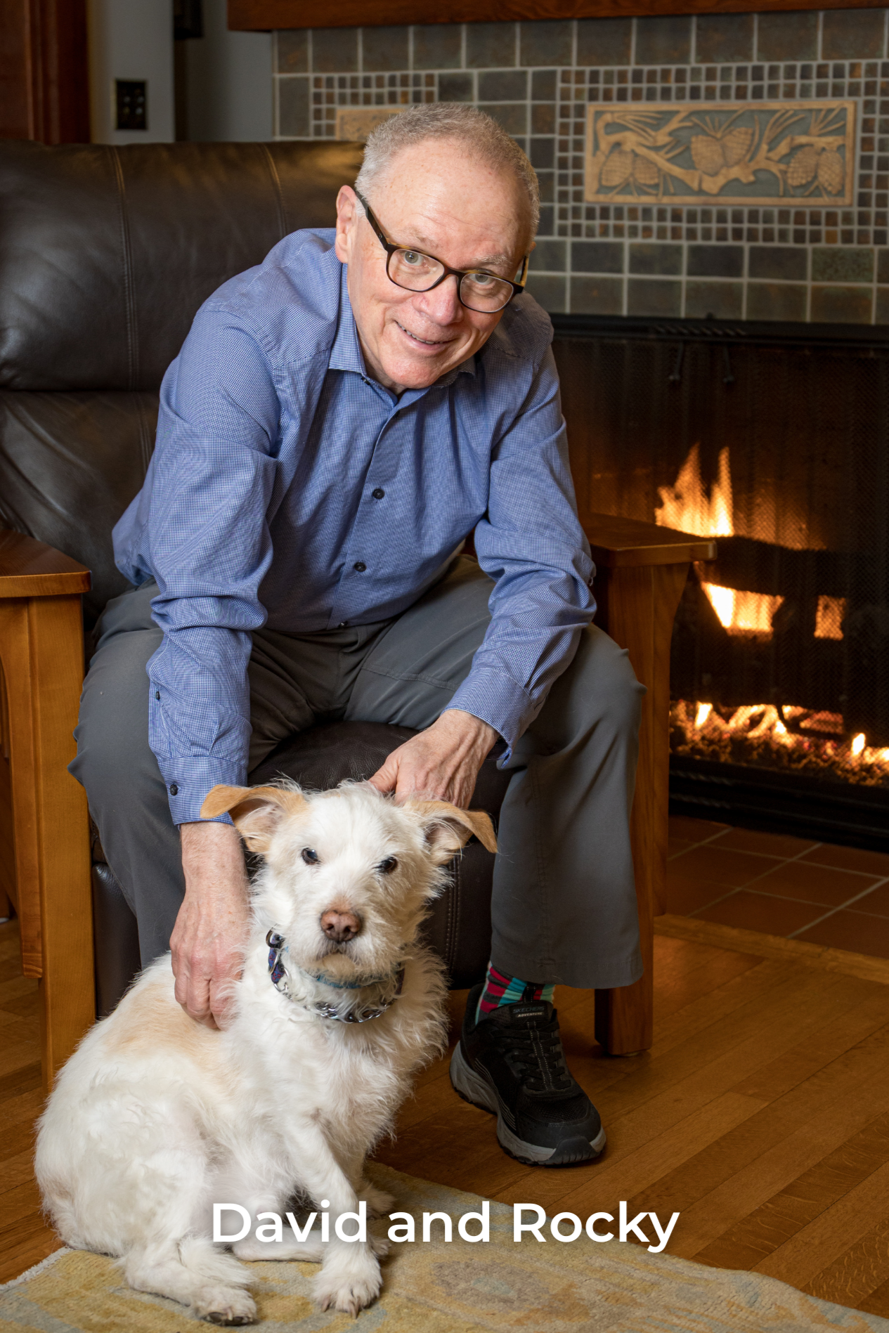 David Womack with glasses in a blue shirt, sitting beside a lit fireplace, gently petting a fluffy, white dog with a collar, sitting on a rug. Text overlay reads "David and Rocky."