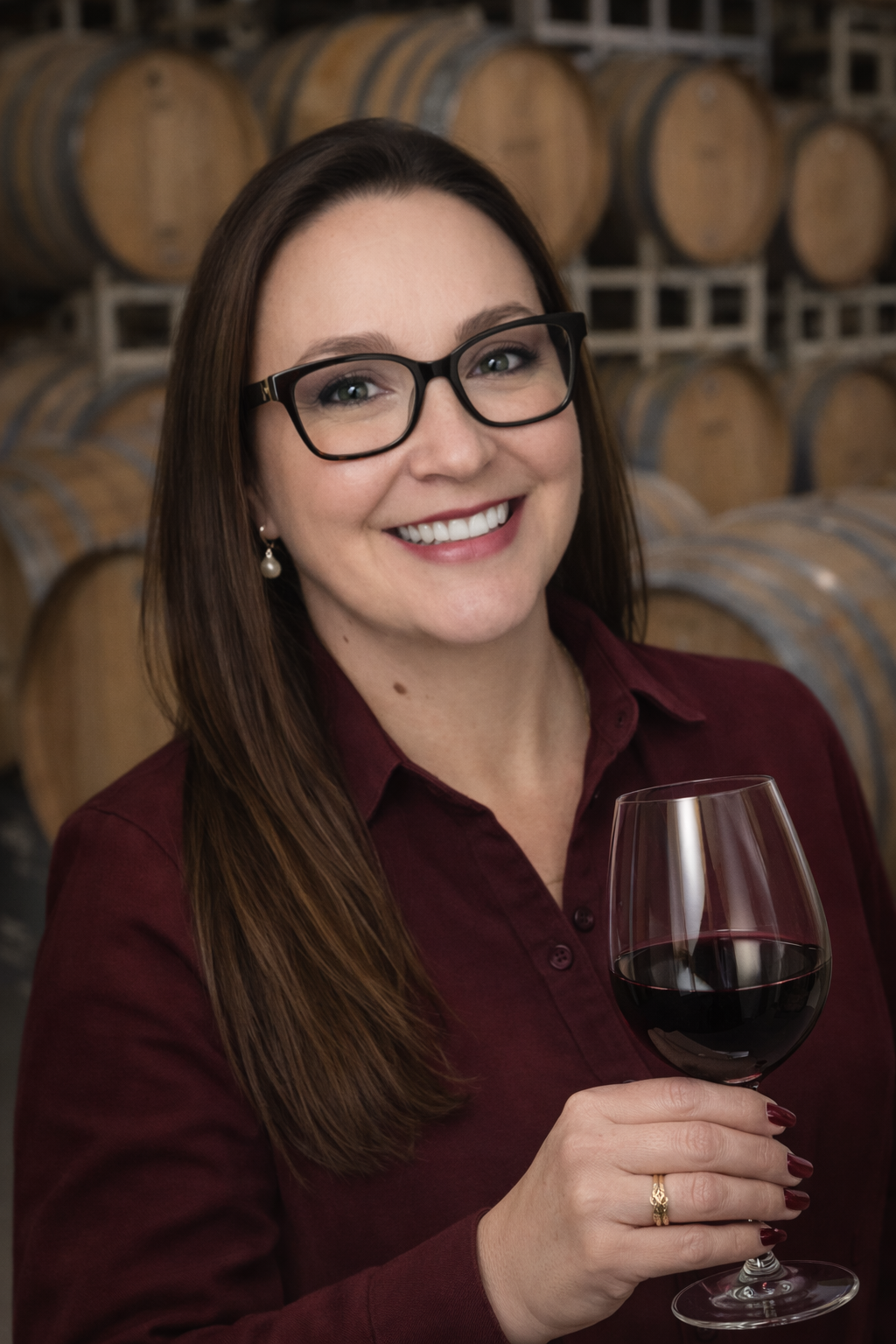 A woman with brown hair wearing glasses and a maroon shirt, holding a glass of red wine, smiling in a winery with wine barrels in the background.