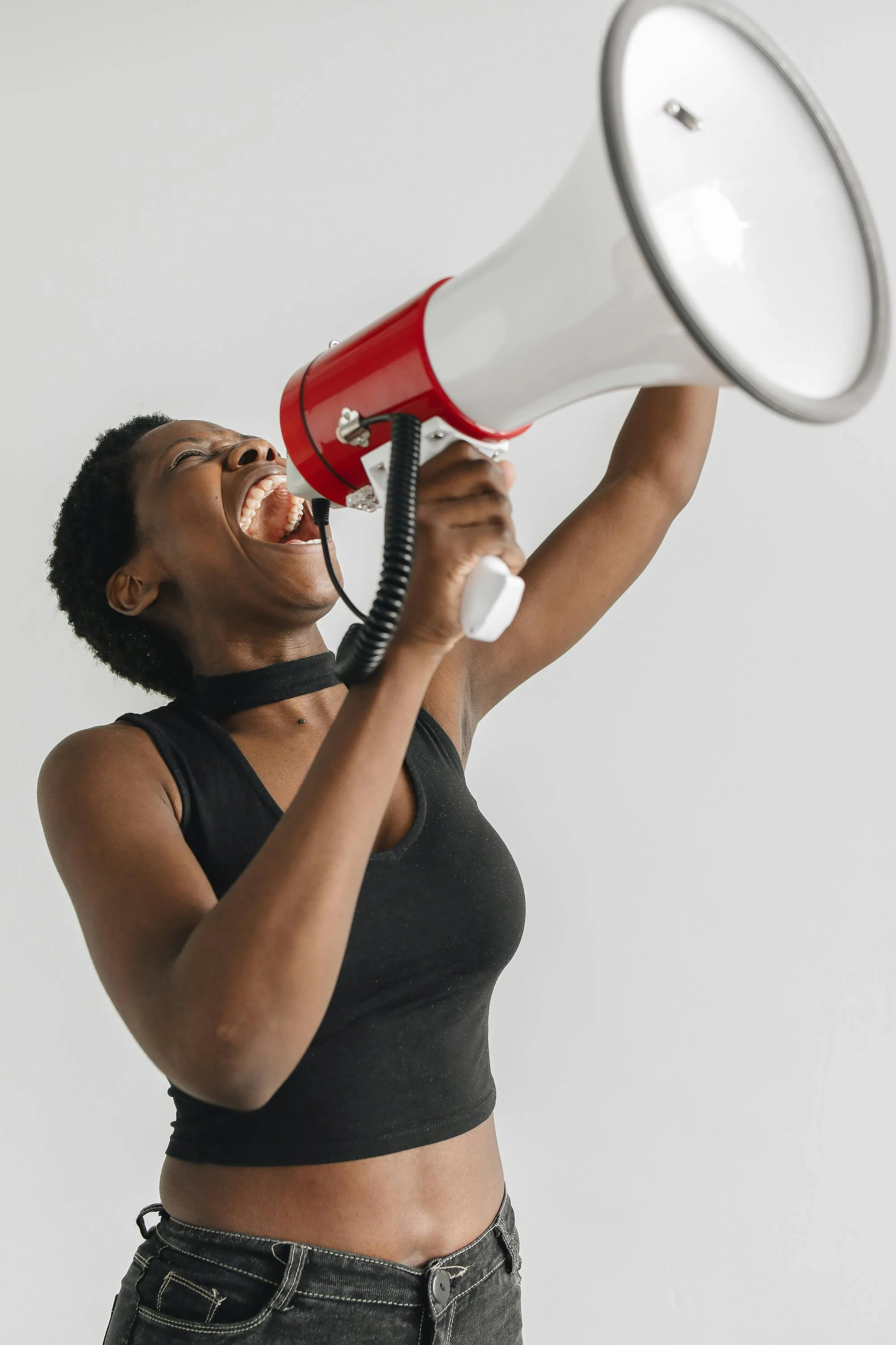 An energetic woman in a black sleeveless top with a choker, holding a red and white megaphone, shouting passionately.