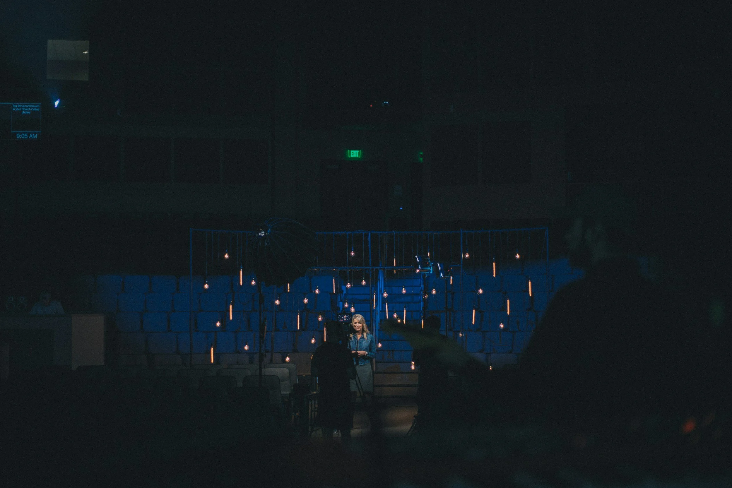 A woman standing in a dark room being filmed or photographed with professional lighting and equipment, with blue chairs and hanging light bulbs in the background.