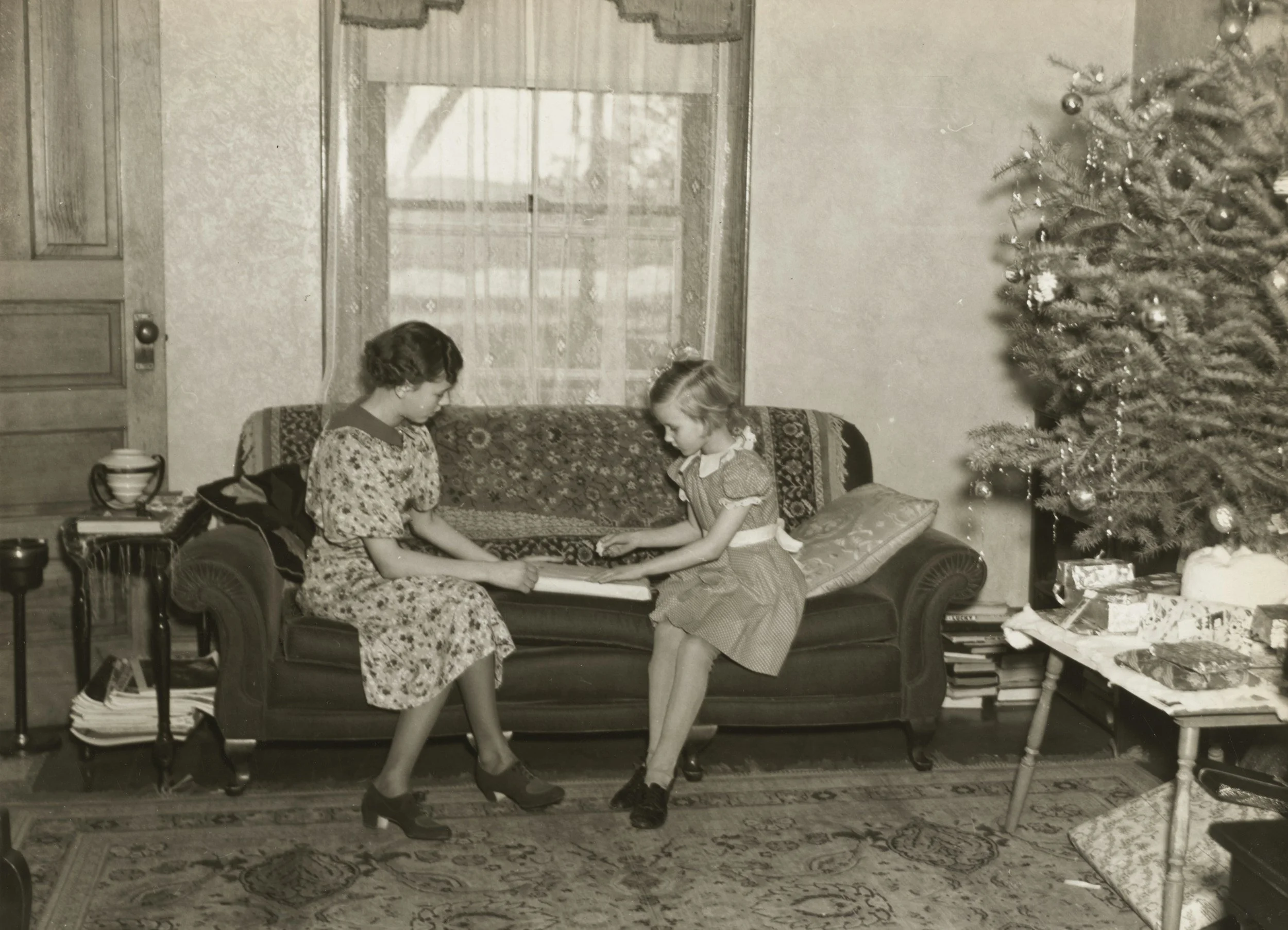 Two young girls in dresses sitting on a vintage sofa, holding hands, in a room decorated for Christmas with a tree and wrapped gifts nearby.