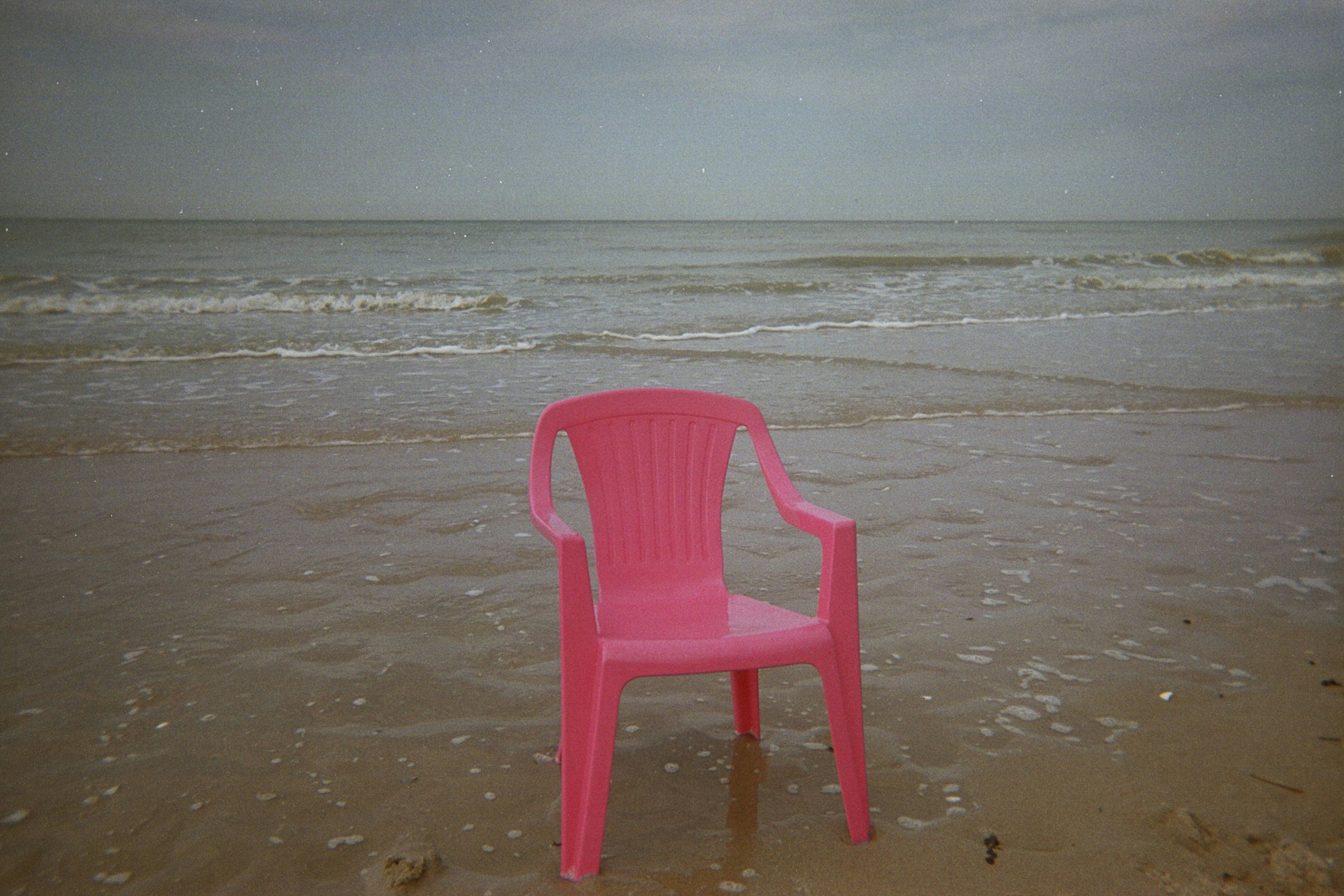 Pink plastic chair on a sandy beach near the ocean, with waves in the background.