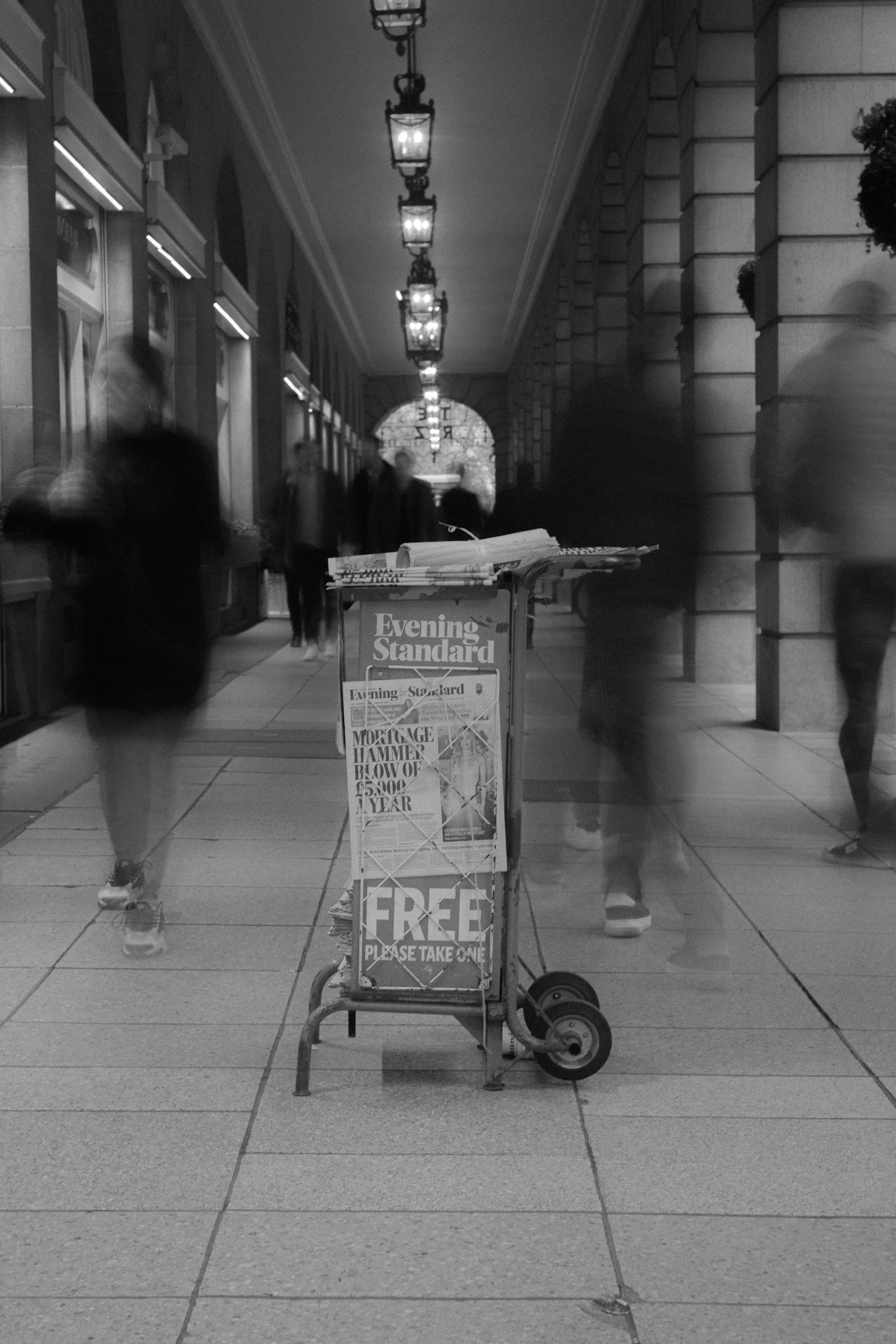 A newspaper stand on a city sidewalk with people walking by in motion blur, illuminated hanging lanterns overhead, and a brick building in the background.