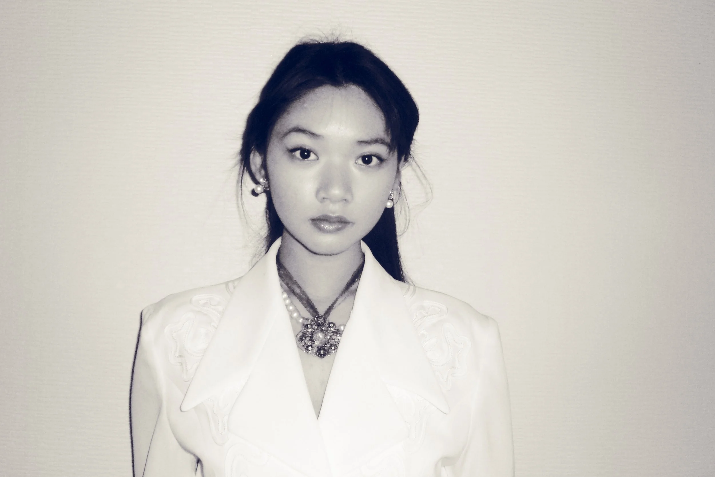A black and white photograph of a young woman wearing a white dress with embroidered details, pearl earrings, and a statement necklace, standing against a plain light-colored wall.