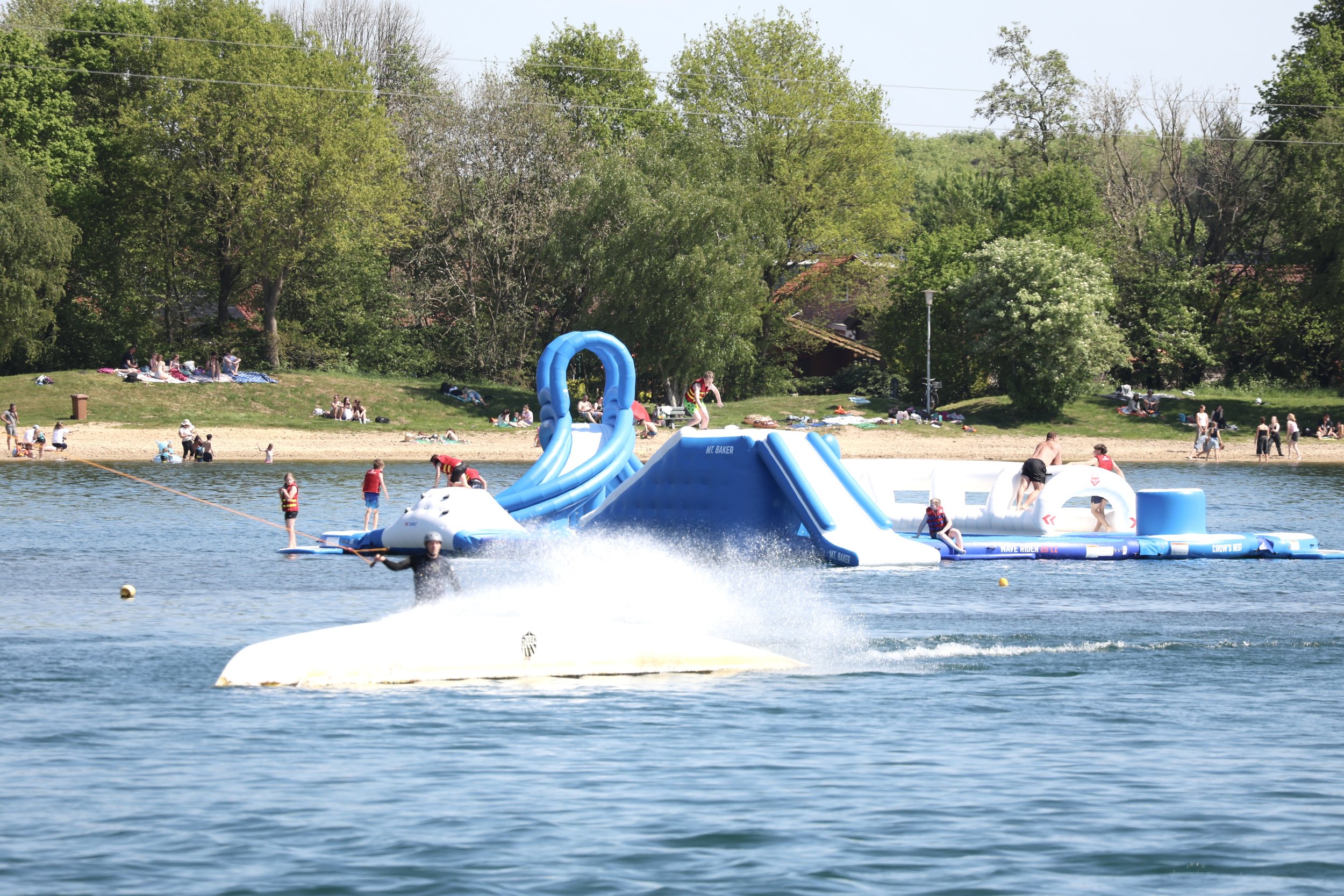 Menschen, die auf einem Wasser-Spielfeld mit Wasser-Rutsche und -Balken spielen, während andere am Ufer entspannen.