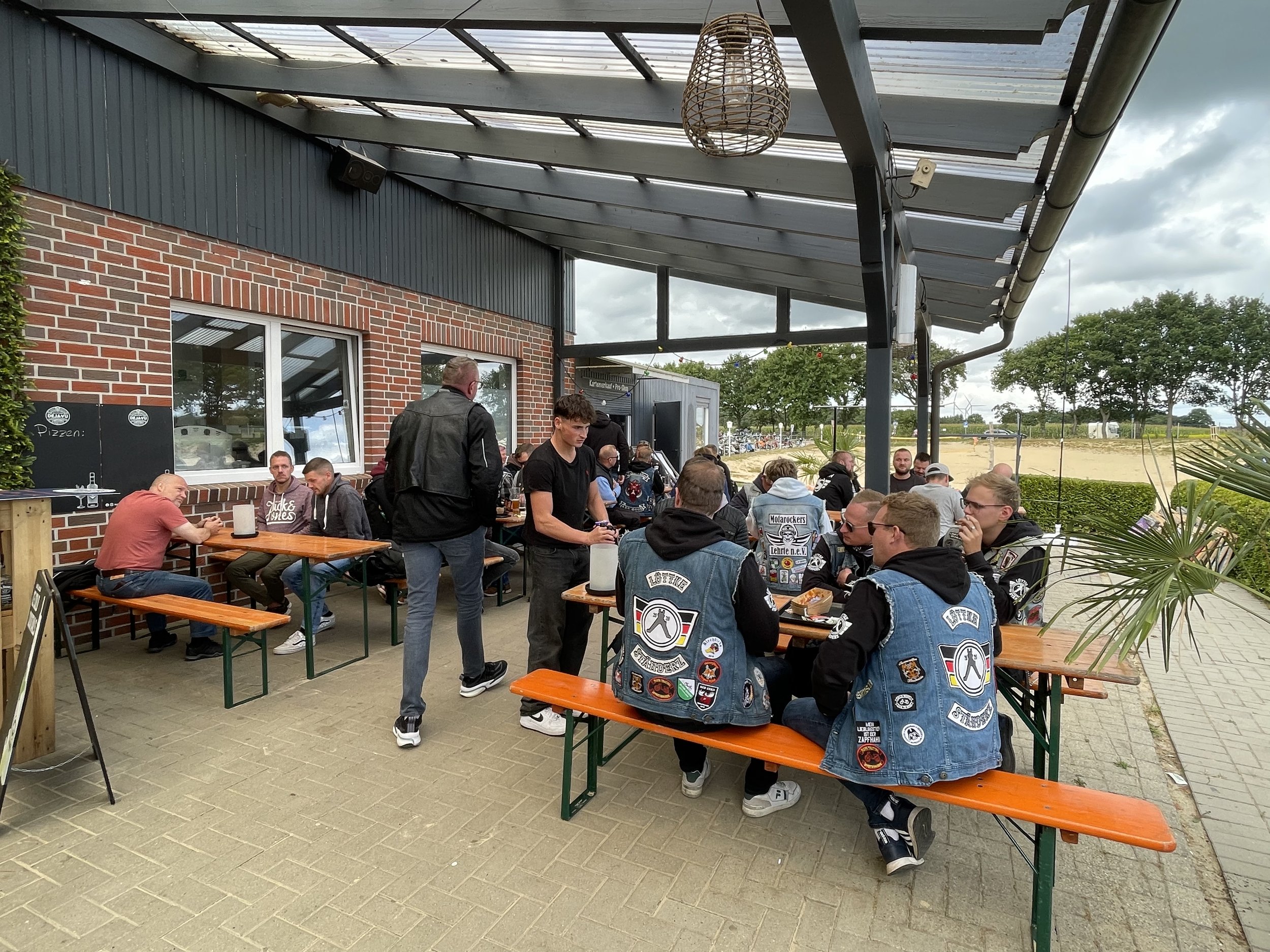Mehrere junge Männer, hauptsächlich Biker mit Jeanswesten mit Aufnähern, sitzen und stehen an Tischen auf einer Terrasse im Freien, die von einem Dach geschützt ist. Im Hintergrund sind Bäume und eine offizielle Sandfläche zu sehen.