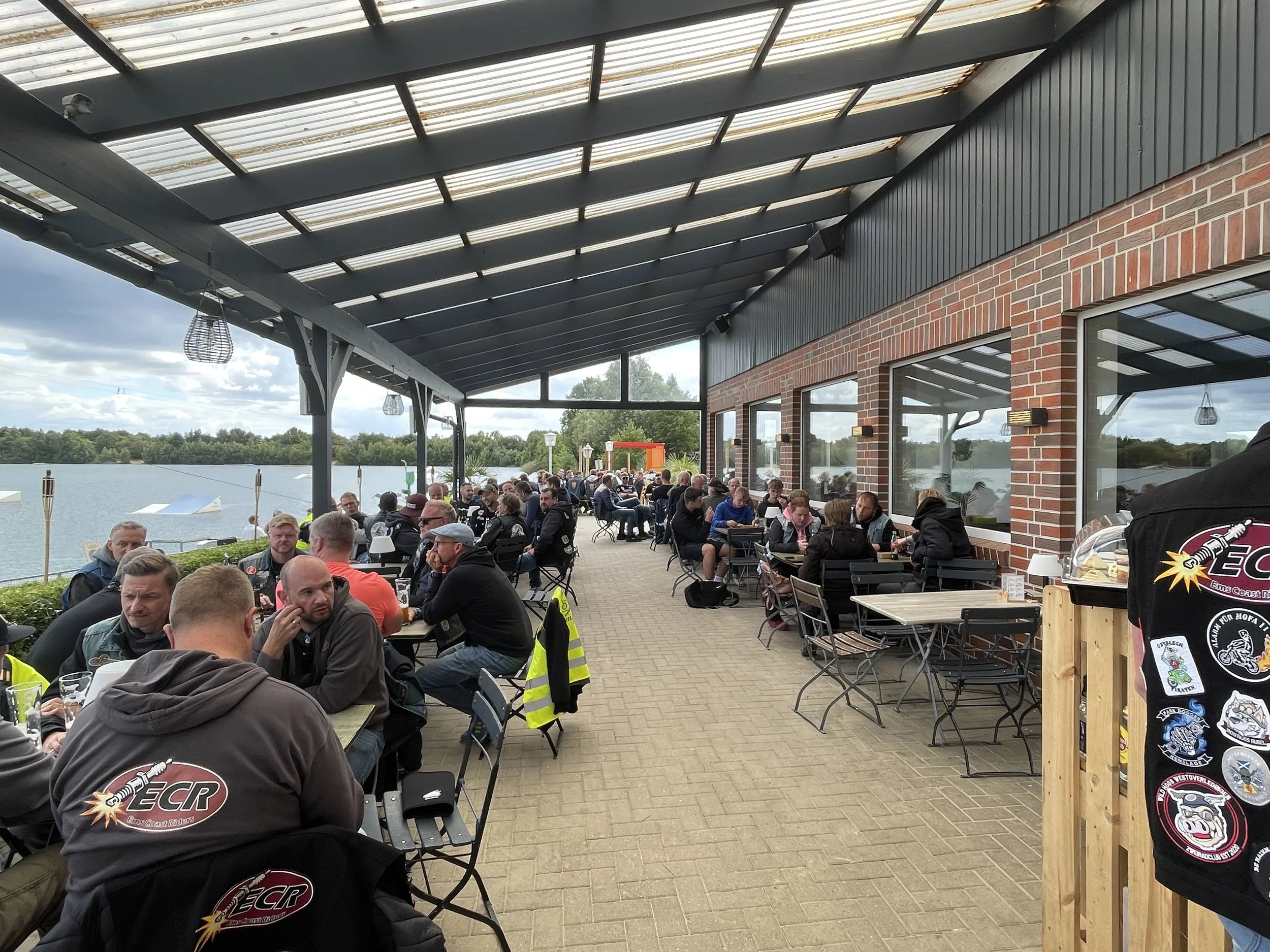 Viele Menschen sitzen an Tischen auf einer Terrasse neben einem Gewässer, blauer Himmel im Hintergrund.