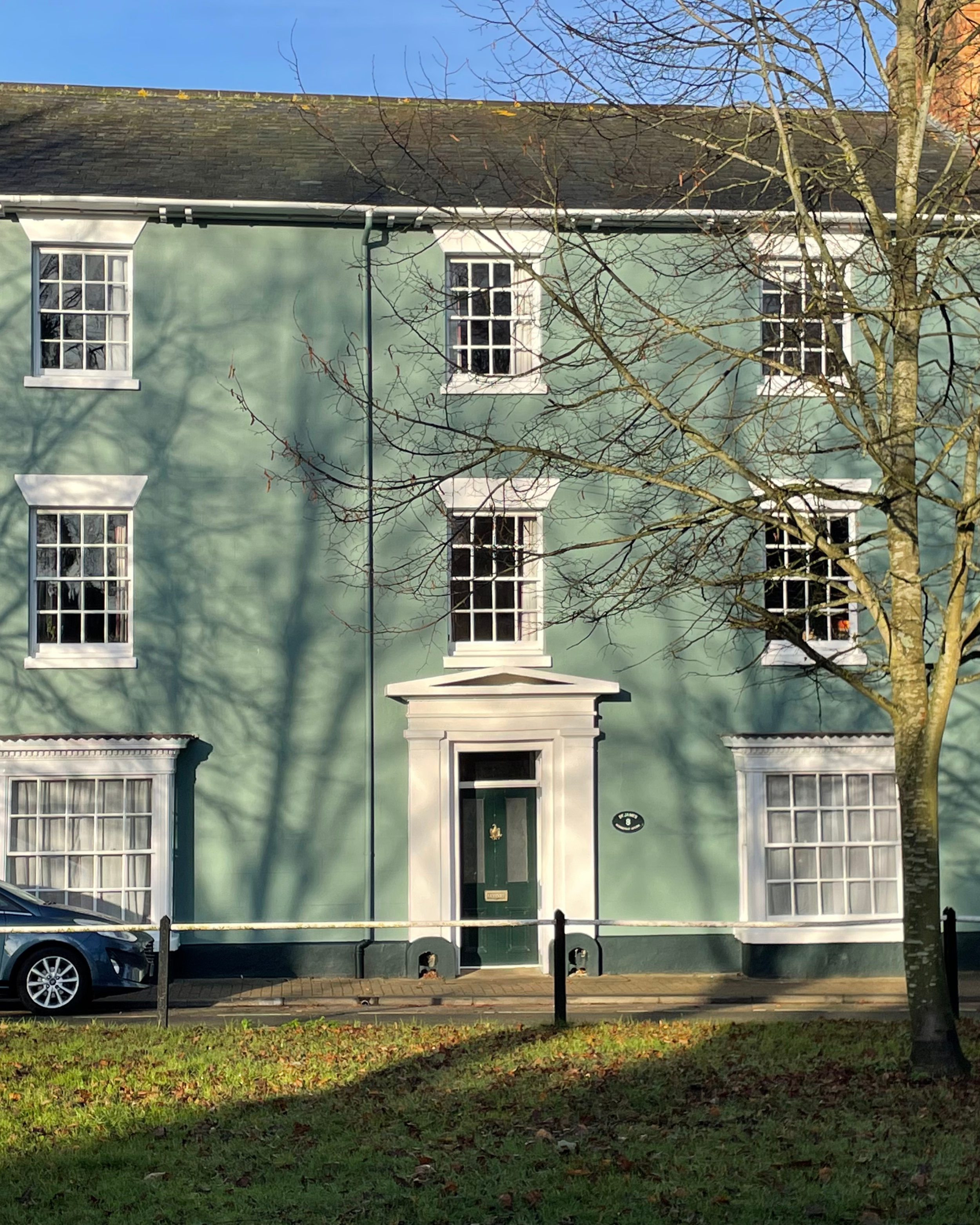 A green, three-story residential building with white trim and six windows, three on each floor. A dark green door with a white frame and a brass knocker is centered on the facade, with a white decorative pediment above it. There is a tree with no lea