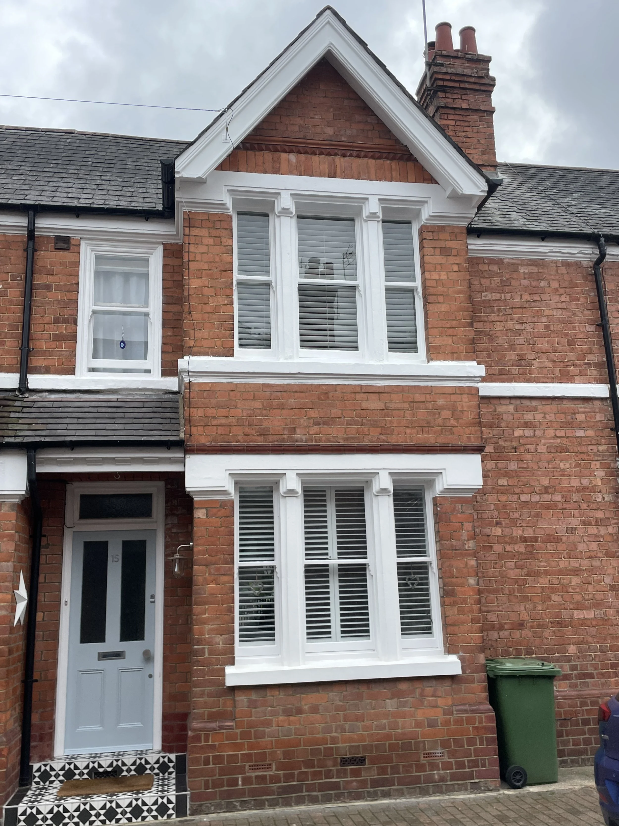 A three-story brick house with white trim and a grey door. The house features large white-framed windows with white plantation shutters. The roof is grey with a chimney on the right side. A green trash bin is placed outside near the house. The sky is