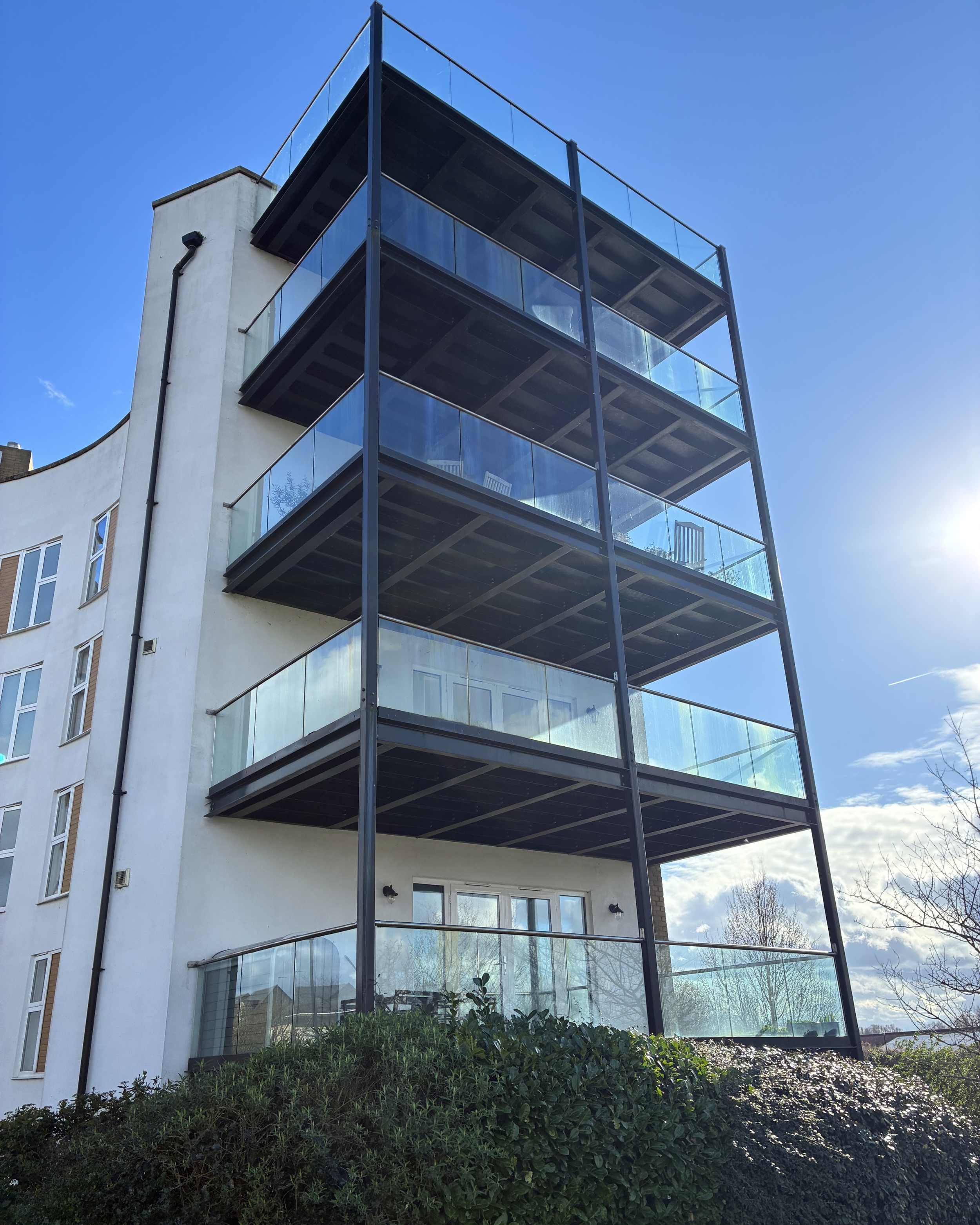 Modern multi-story apartment building with glass balconies and black metal frame, set against a blue sky with sunlight.
