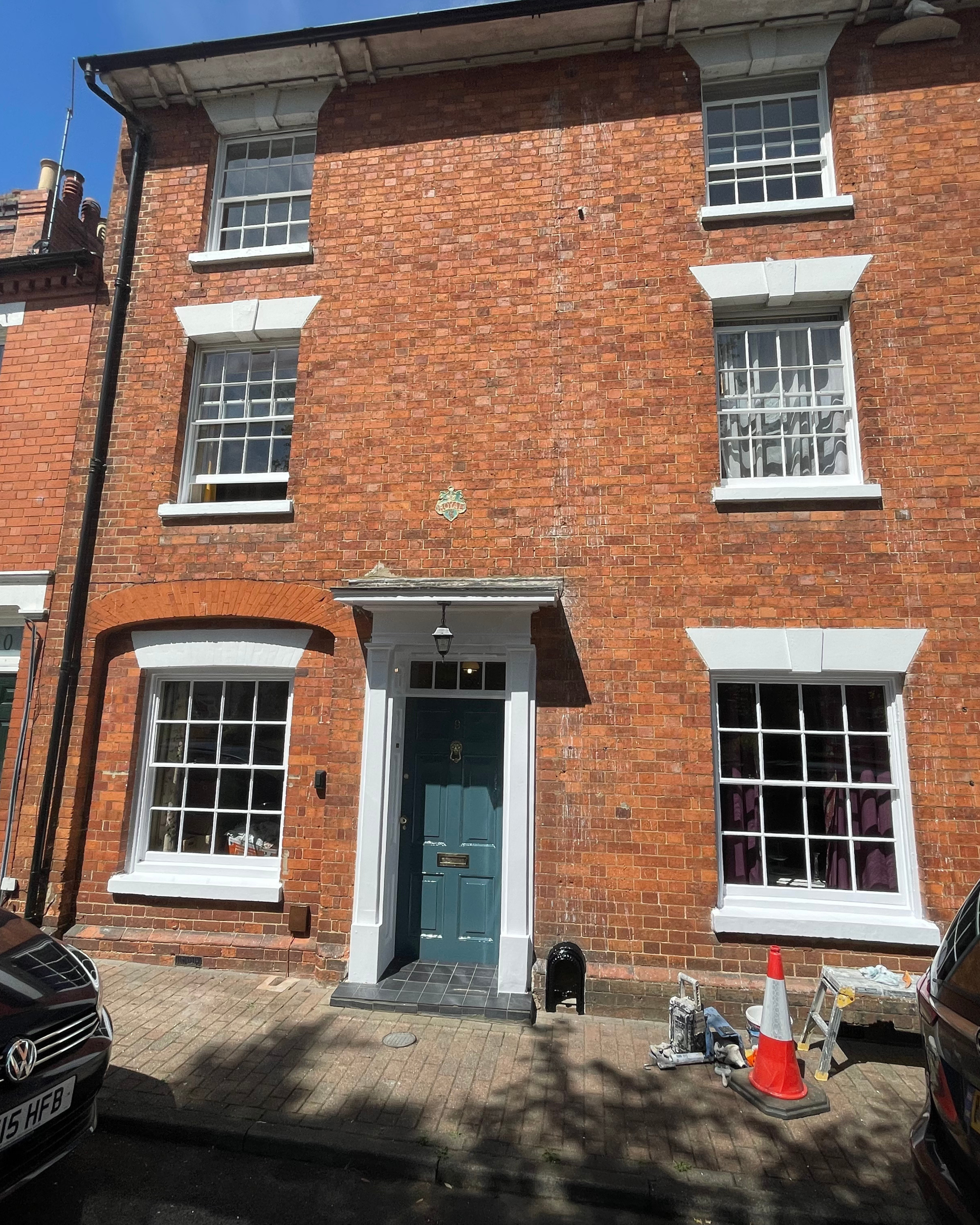 A three-story red brick house with white window trims and a blue front door. There is a small porch with a black hanging light above the door. Construction tools and a traffic cone are in front.