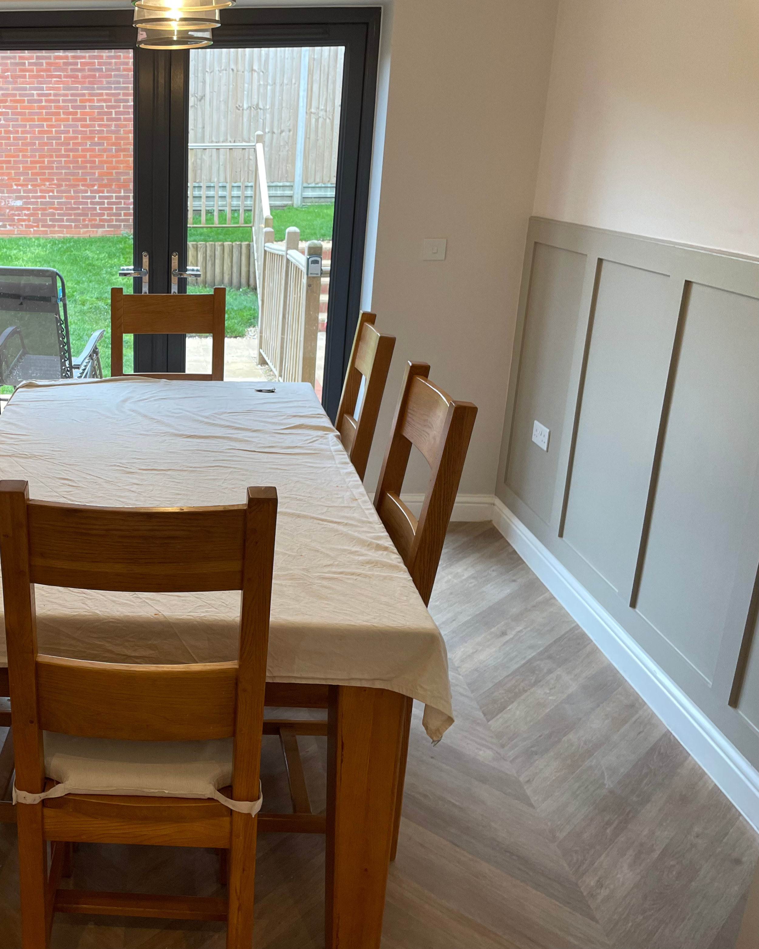 Dining room with a wooden table covered by a white cloth and four wooden chairs. There is a sliding glass door leading to a patio with a small fenced yard and a brick house in the background.