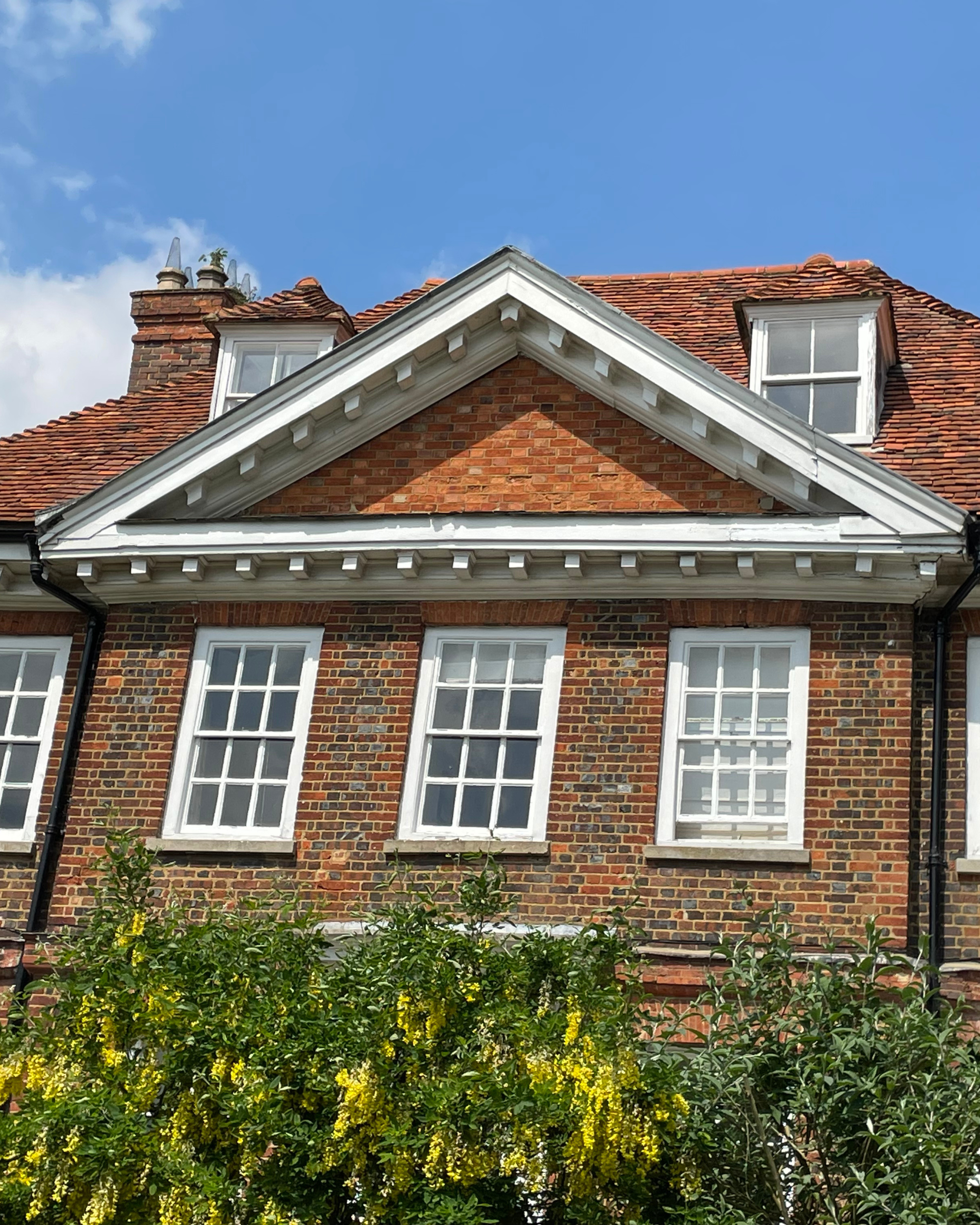 A brick house with six white-framed windows on the front and a triangular roof section with white decorative trim, red clay tile roof, and small dormer windows, with green bush and yellow flowers in the foreground under a partly cloudy blue sky.