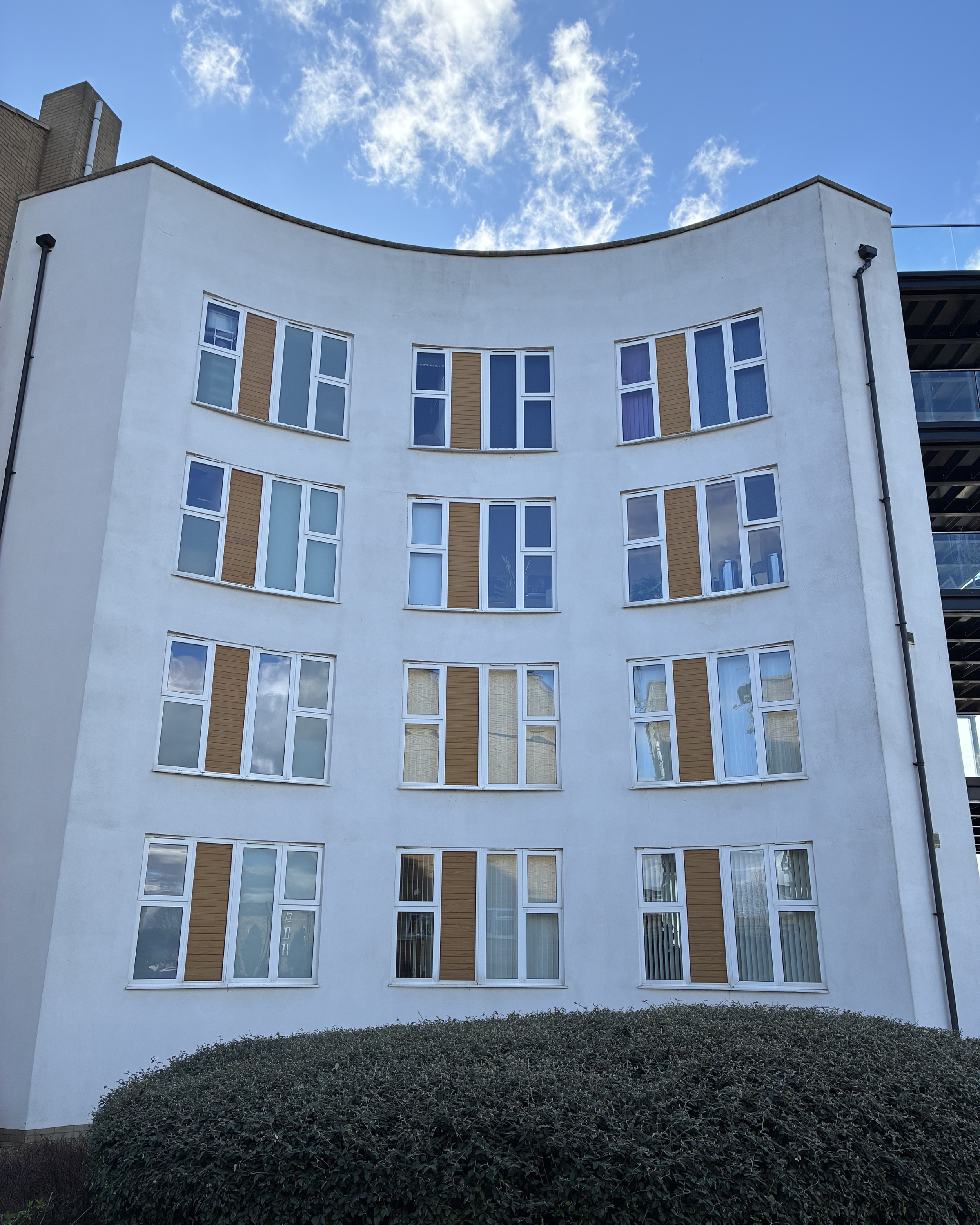 A white, curved apartment building with multiple windows and brown accents under a partly cloudy blue sky.