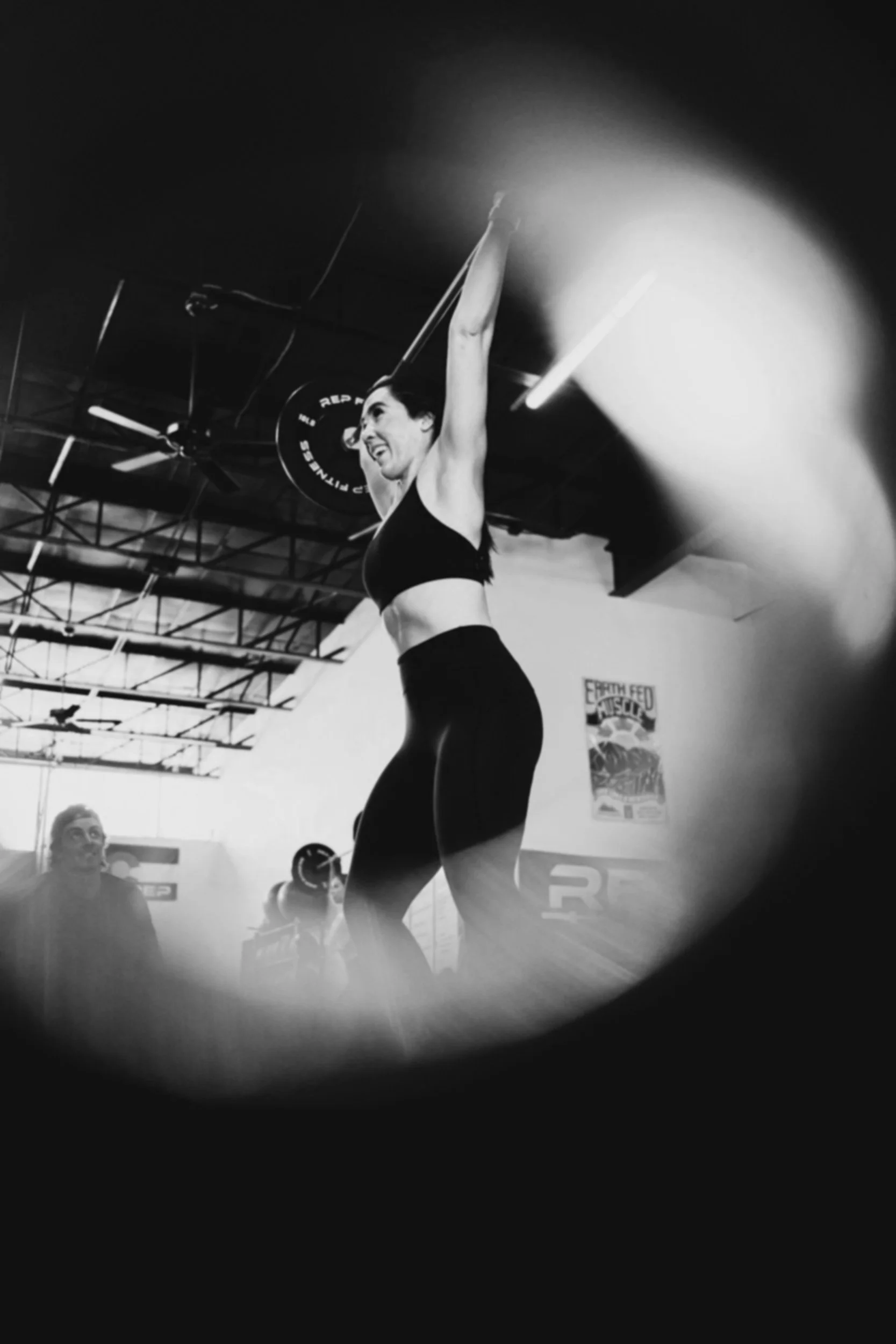 A woman lifting a barbell overhead during a workout at a gym, with a woman in the background observing. Black and white photo with a vignette effect.