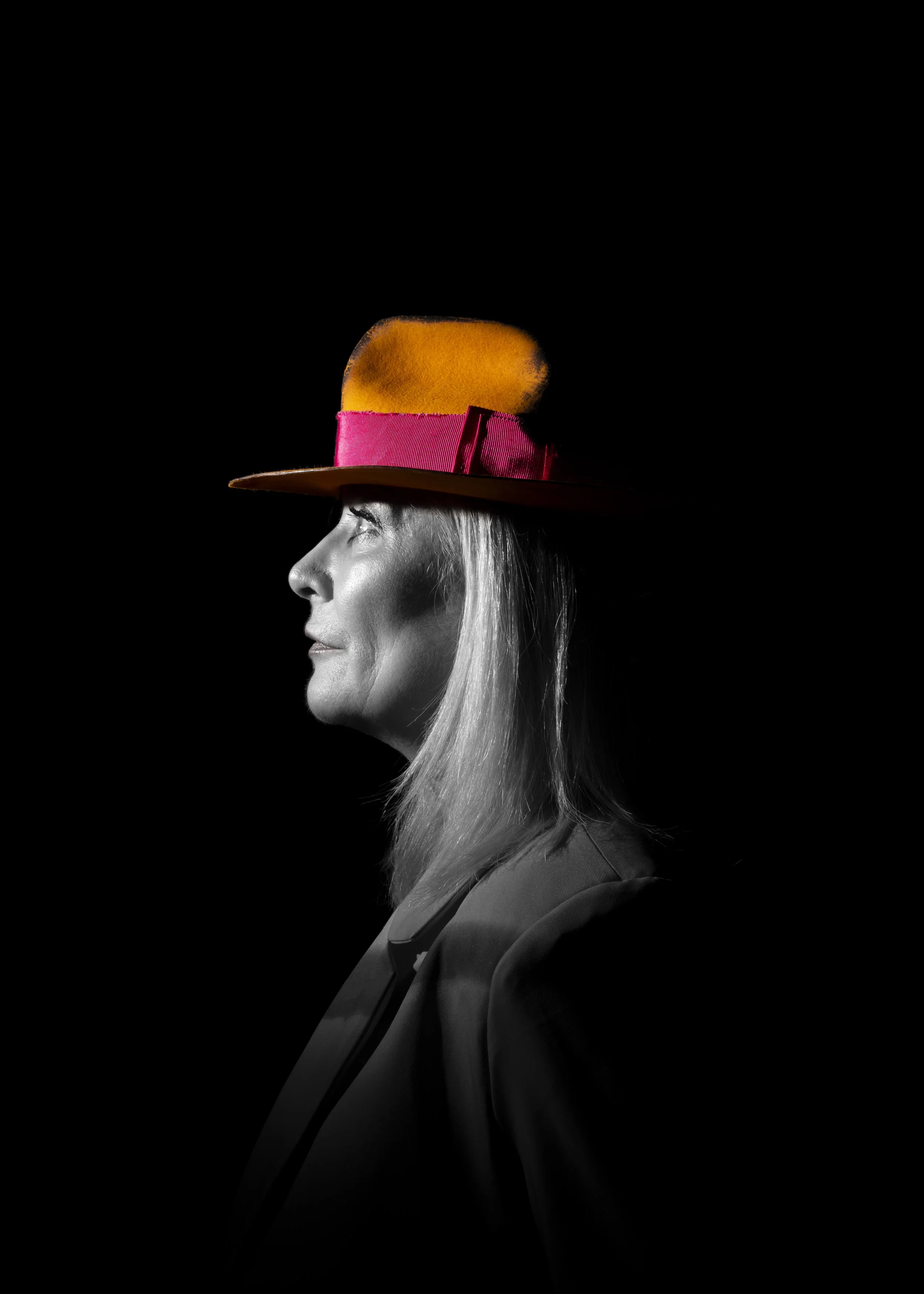 Black and white profile portrait of a woman with an orange hat, featuring a pink band, against a black background.