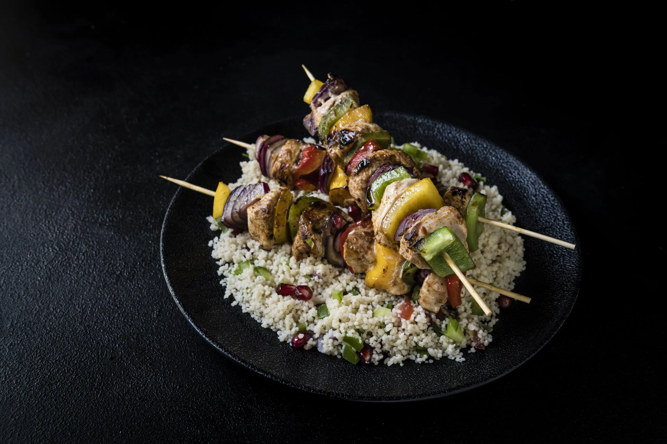 Skewers of grilled chicken and vegetables on a bed of couscous on a black plate.