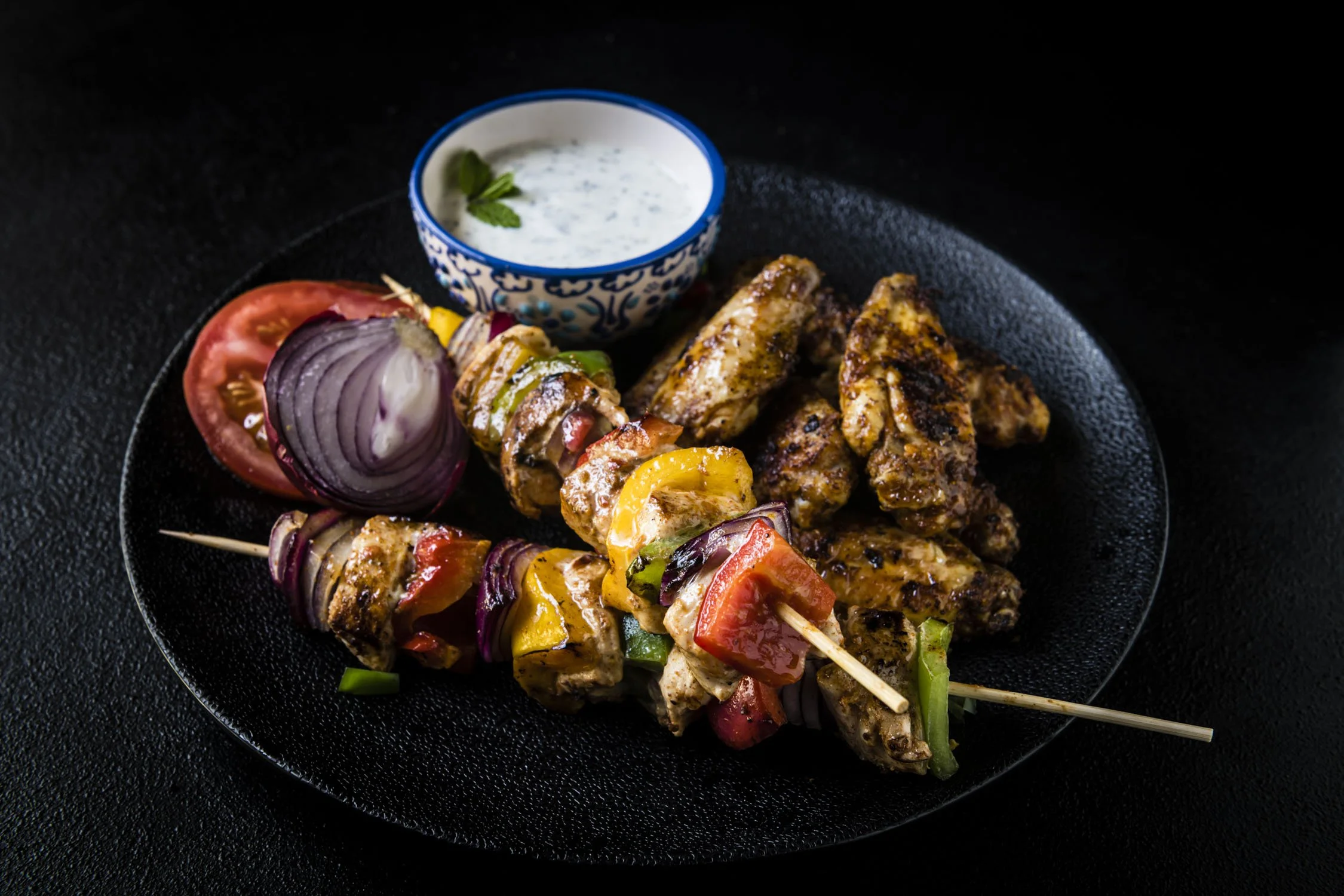 Mixed grilled skewers with vegetables, grilled chicken, sliced tomato, red onion, and a side of creamy herb sauce in a patterned bowl on a black plate.
