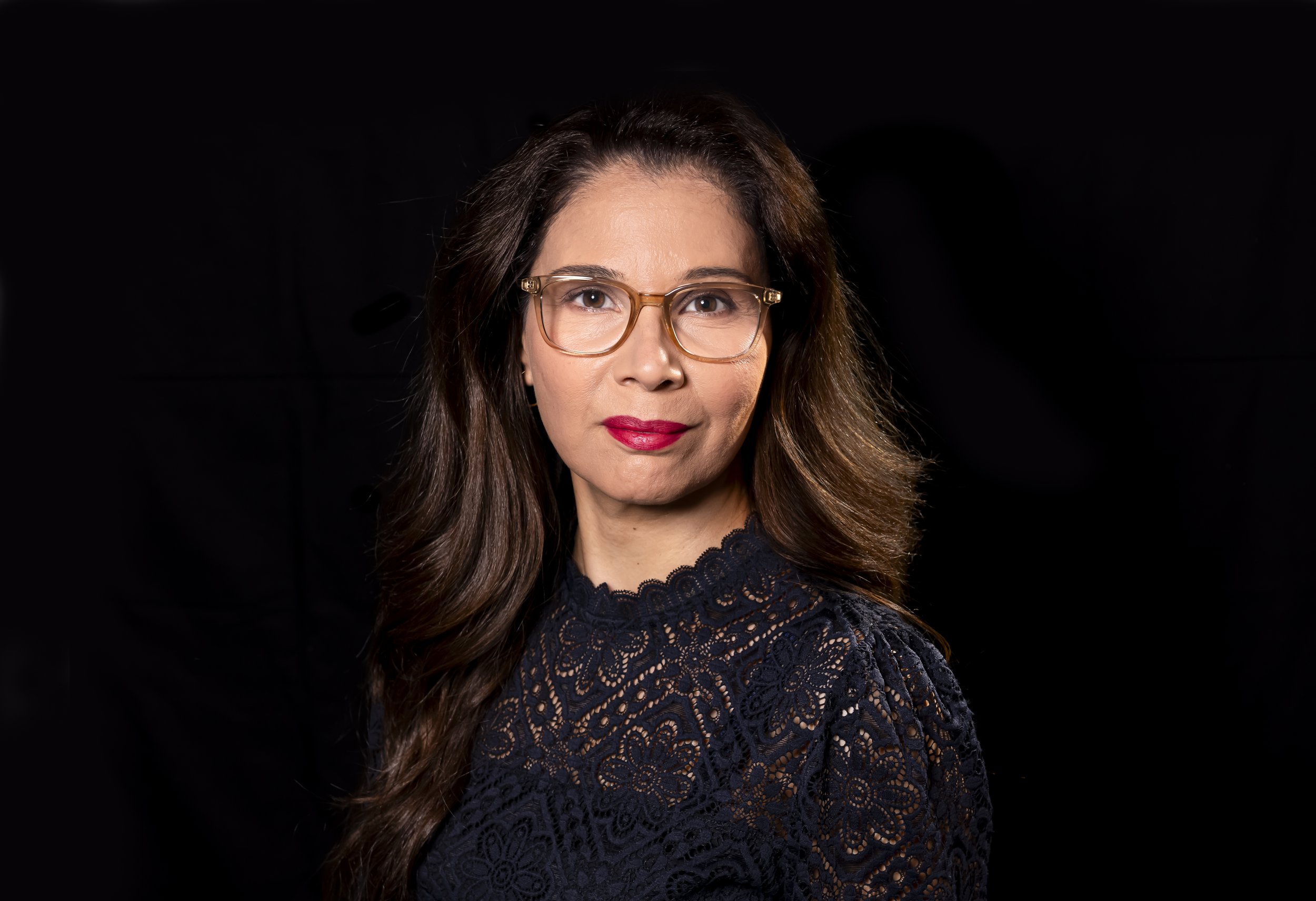 A woman with long brown hair wearing glasses and a dark lace top posing against a black background.