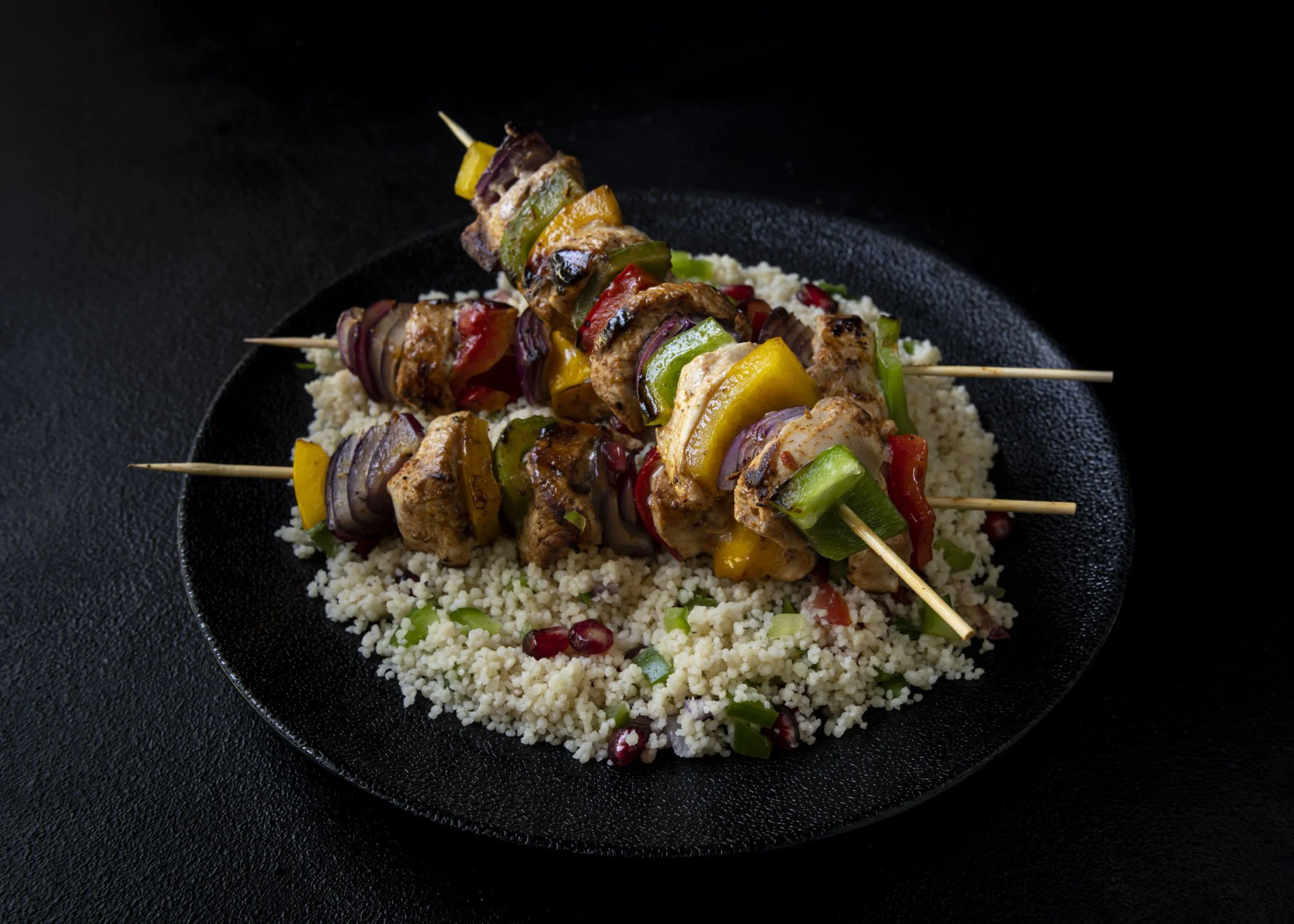 Skewers with grilled meat and vegetables on a bed of couscous, served on a black plate.