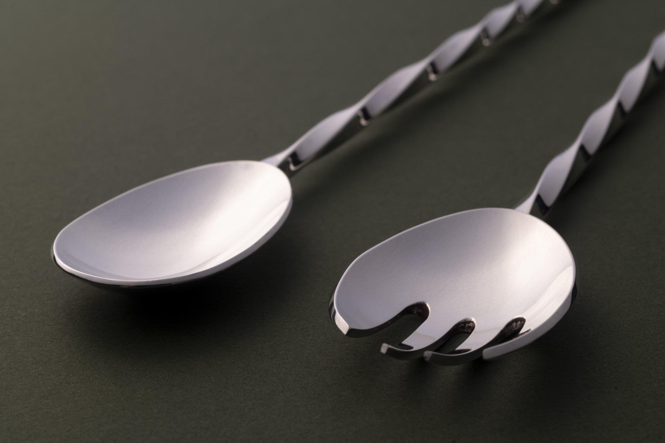 A close up of a silver dessert spoon and fork with elegantly twisted handles on a dark olive green surface.