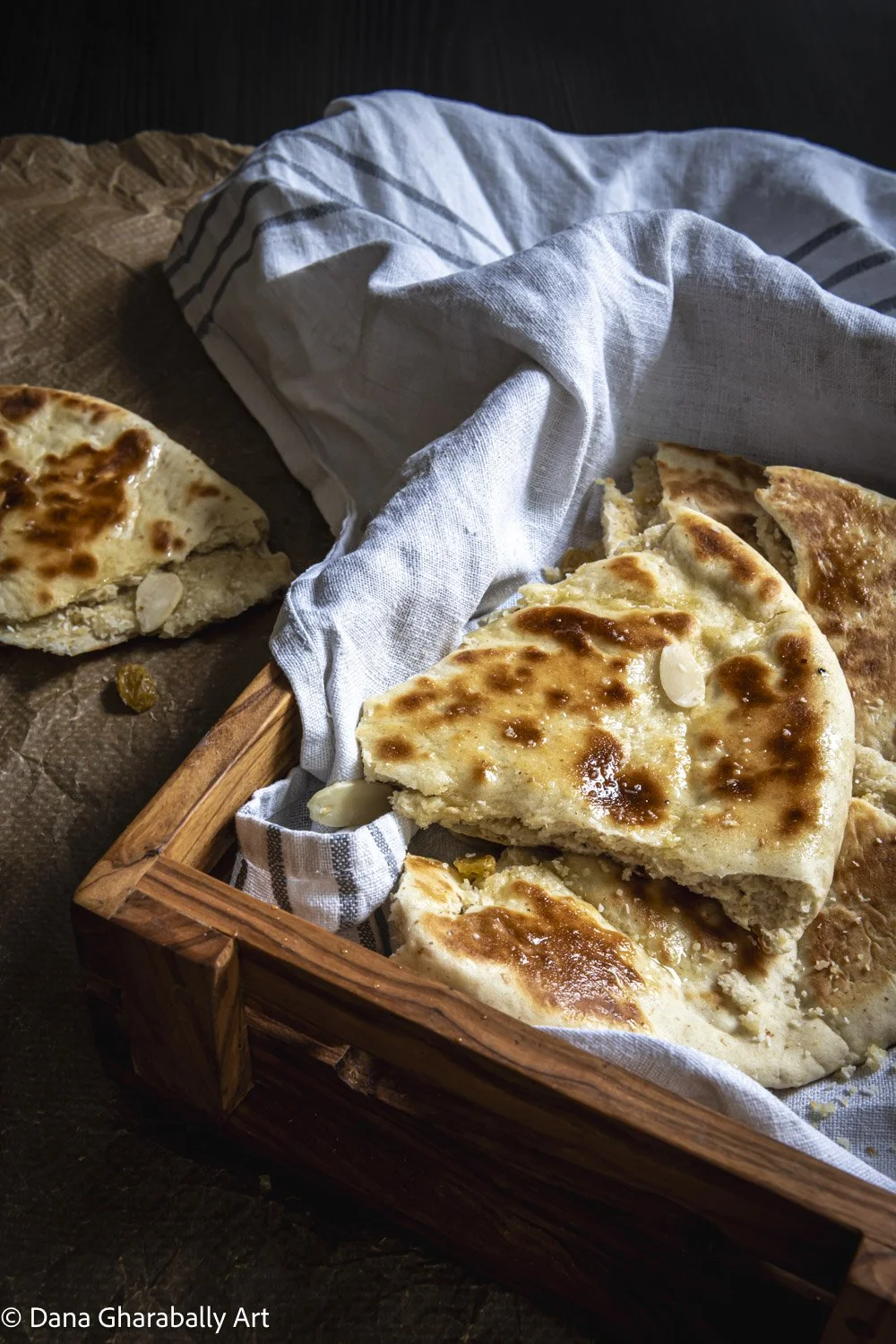 A wooden tray filled with Peshwari Naan bread with a slice and some crumbs on a dark surface, lined with a striped cloth. A piece of bread is on the dark table surface nearby.