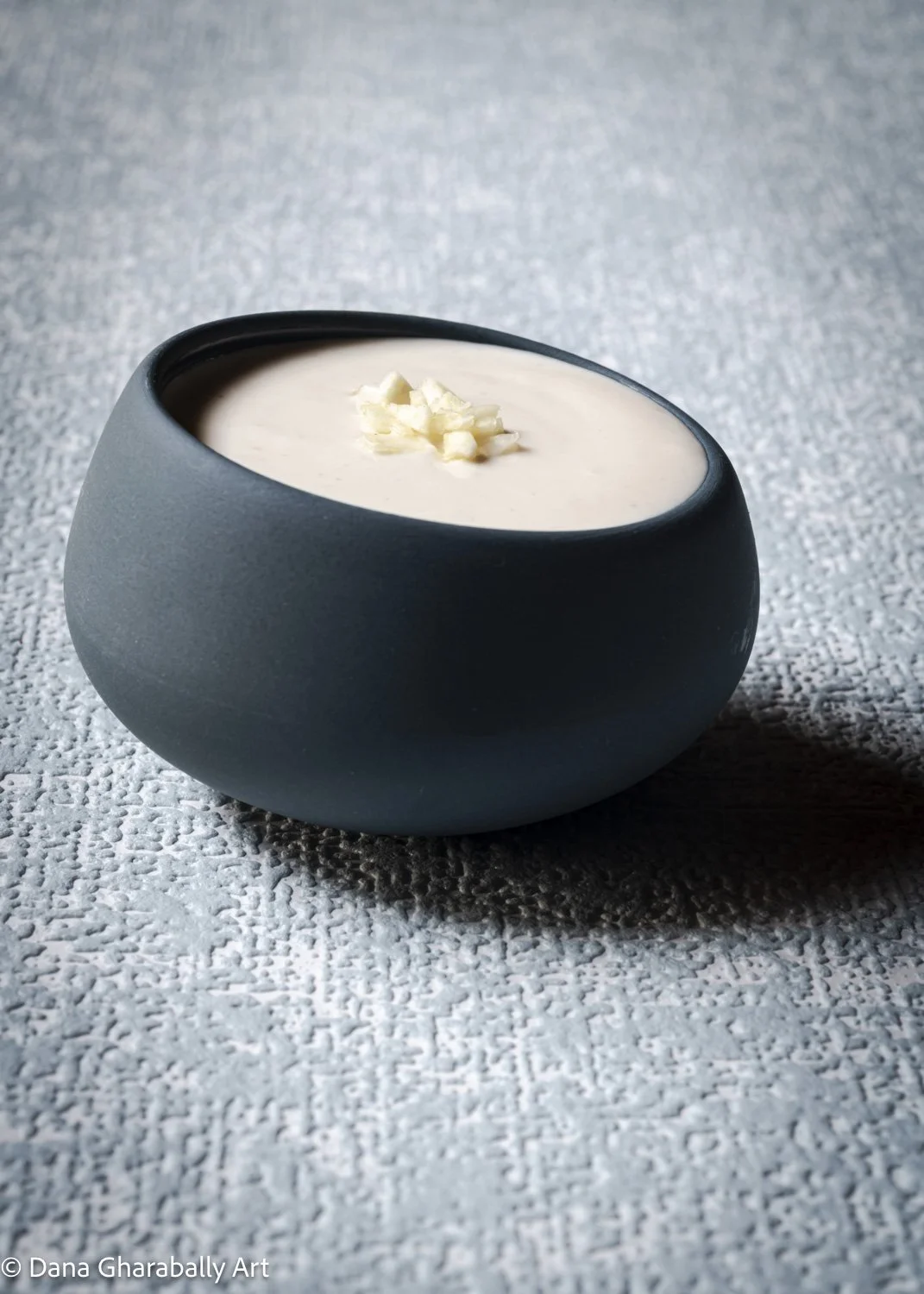A blue bowl filled with white creamy garlic dip topped with finely diced fresh garlic. The bowl sits on a textured surface.