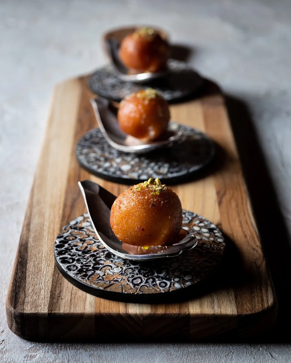 Three Gulab Jamun, each topped with crushed pistachios, served on decorative spoons placed on a wooden board.