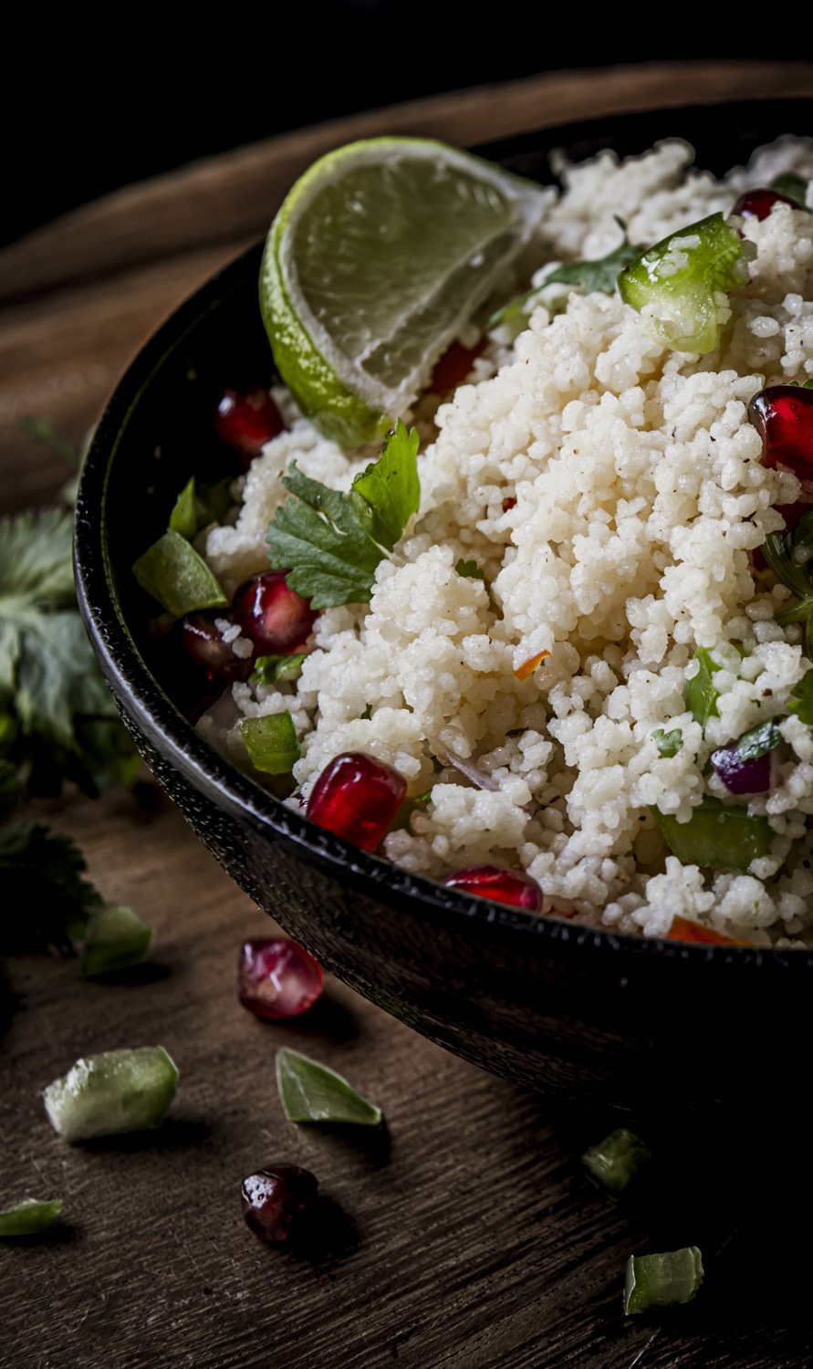 A bowl of couscous salad garnished with pomegranate seeds, chopped green vegetables, coriander, and a lime wedge, placed on a wooden surface.