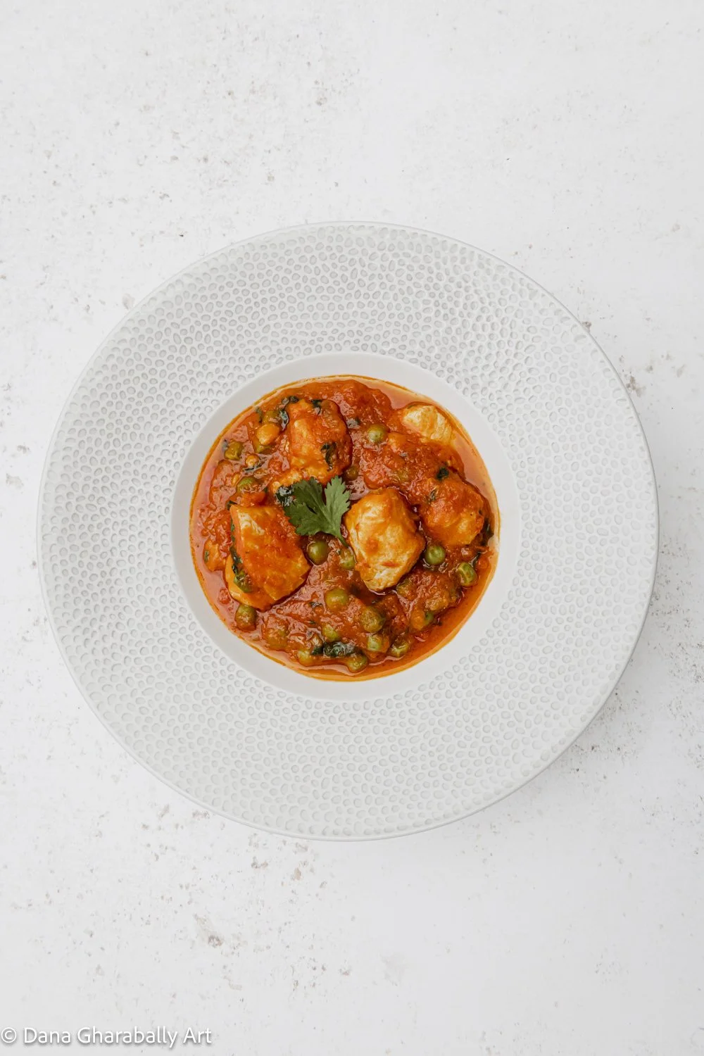 Bowl of fish curry with peas garnished with coriander on a white textured plate.