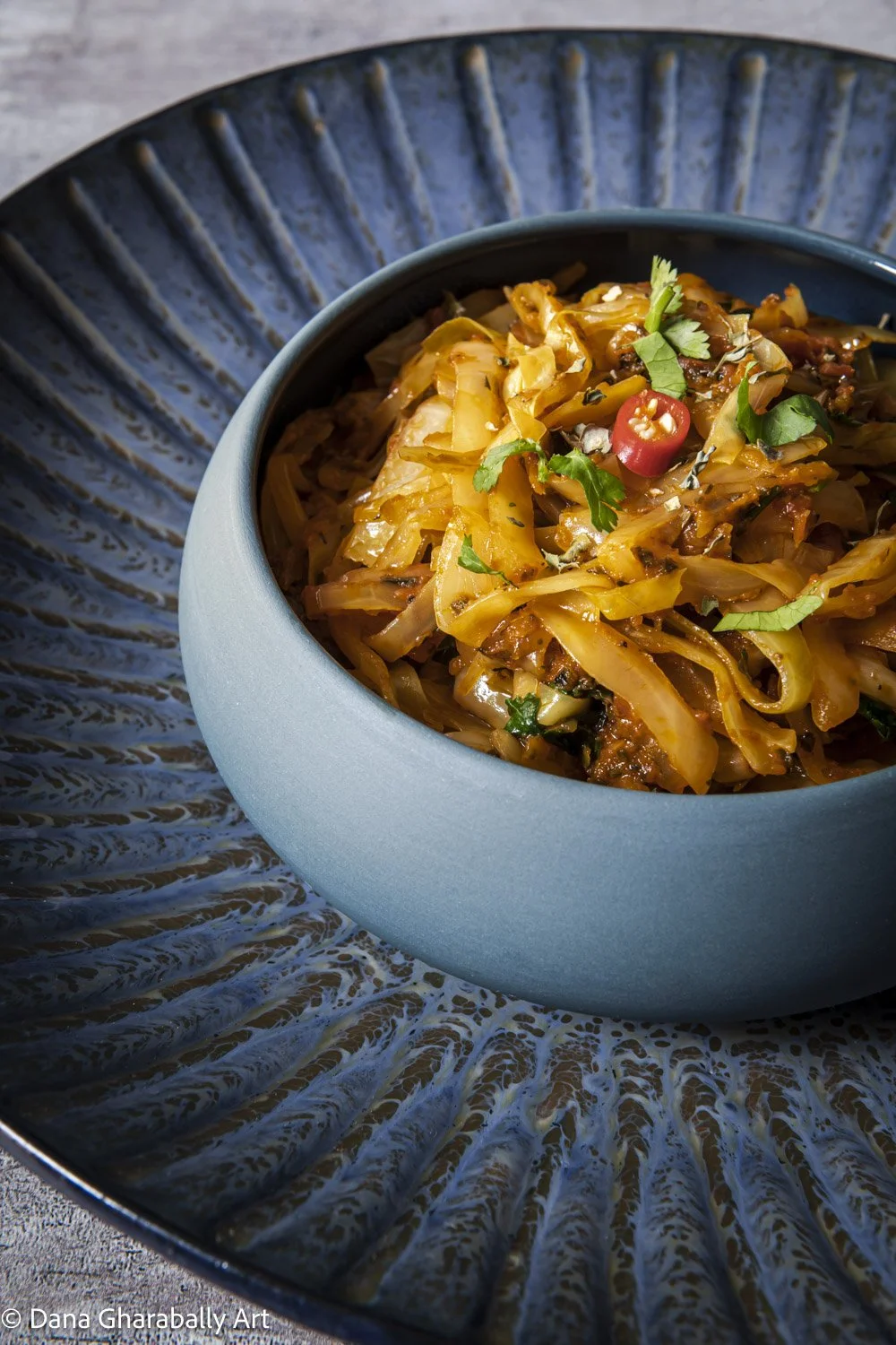 A blue bowl filled with cooked stir-fried cabbage, garnished with fresh coriander and a sliced red chilli, on a decorative round blue plate.