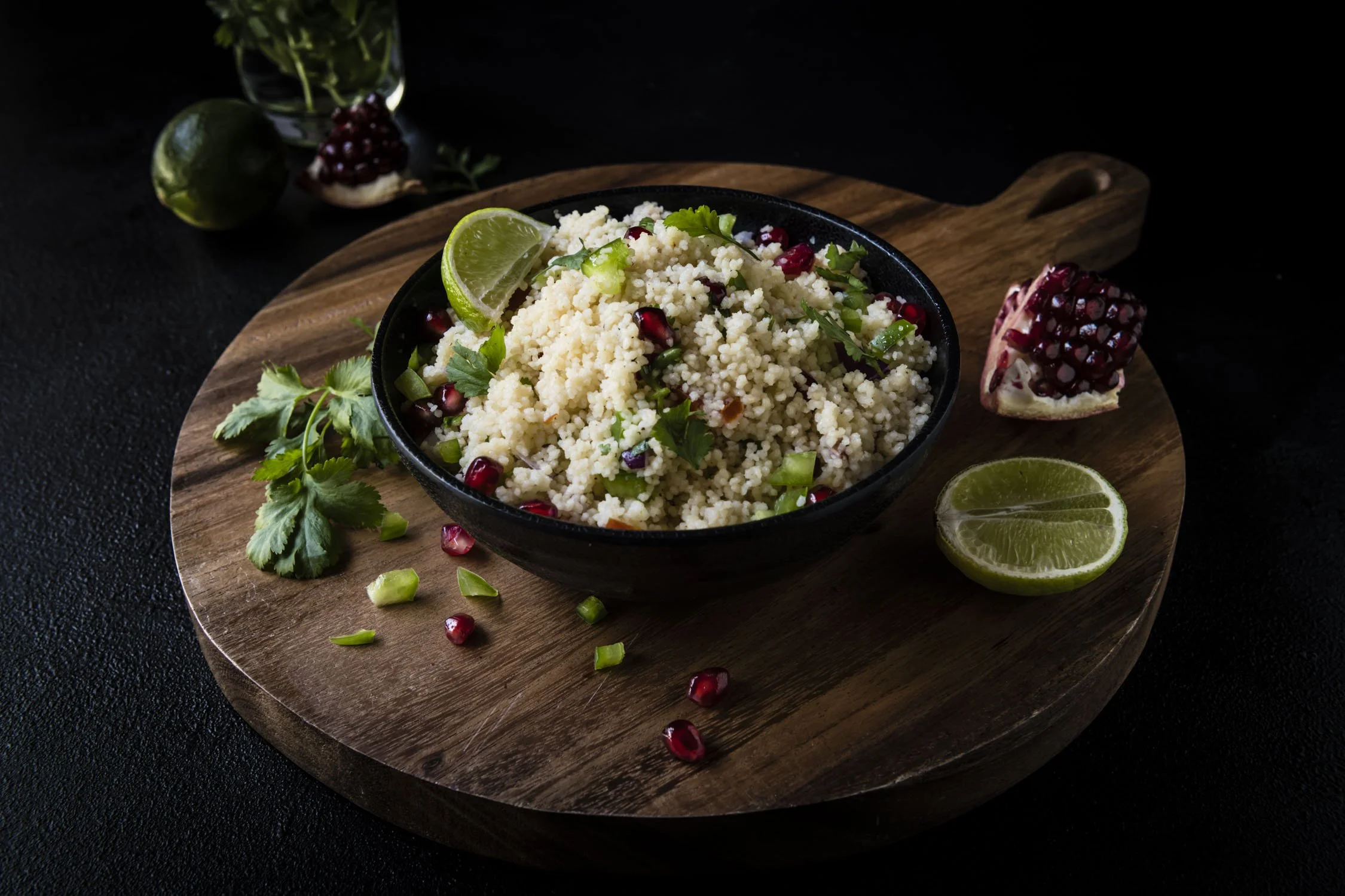 A black bowl of couscous garnished with pomegranate seeds, chopped green onions, coriander, and lime wedges, set on a round wooden cutting board with pomegranate halves and a lime wedge nearby.