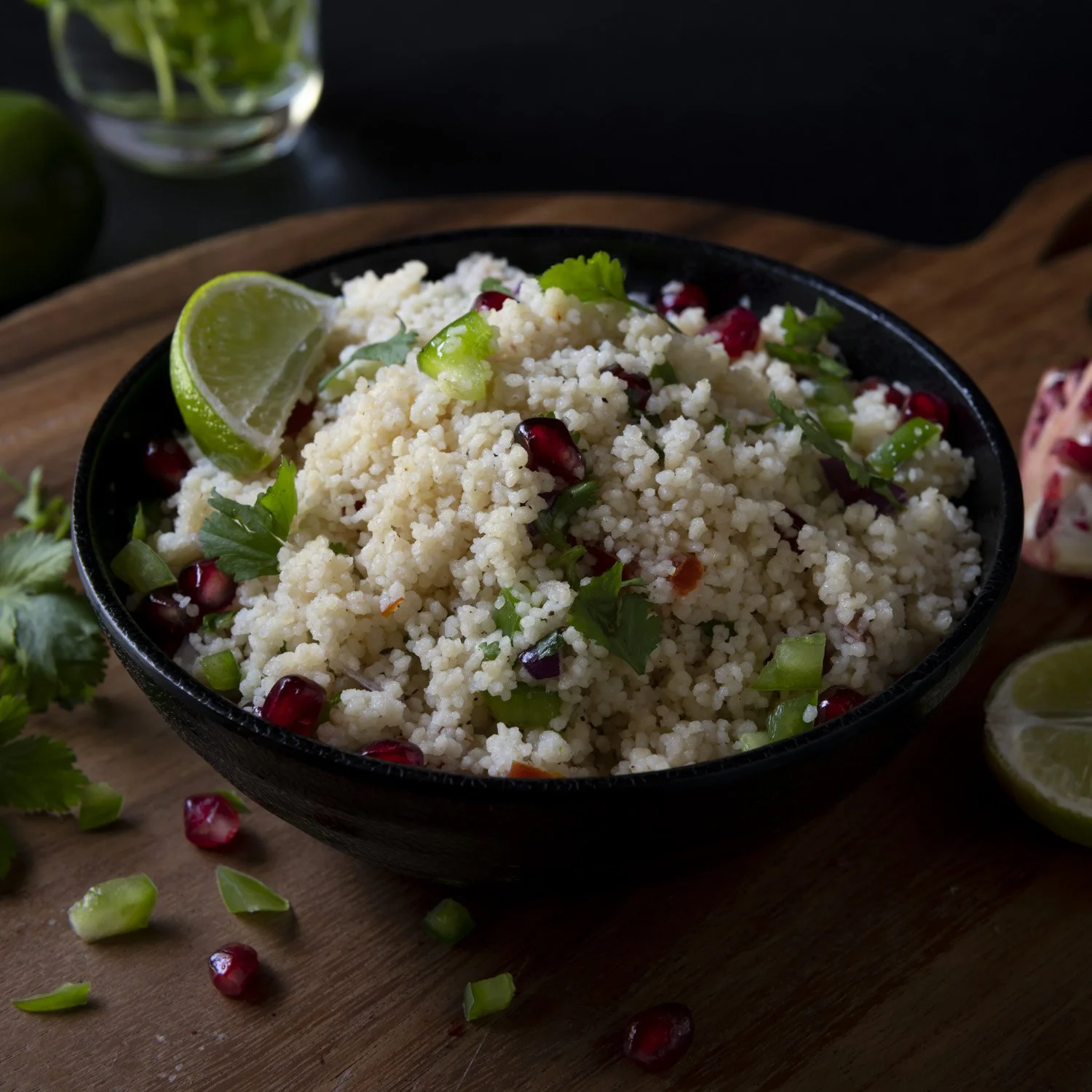 Bowl of couscous salad with pomegranate seeds, chopped green onions, coriander, and lime wedge on a wooden board.