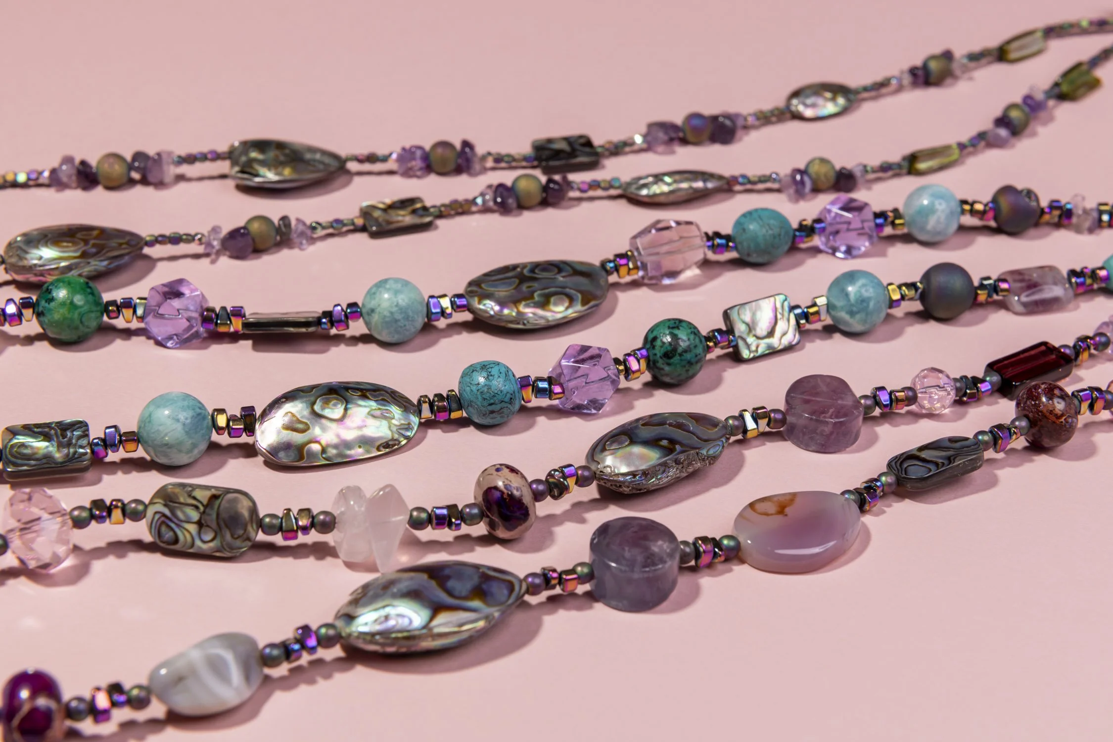 Multiple strands of colourful beaded necklaces with various beads including shell, glass, and stone, laid out on a pink background.