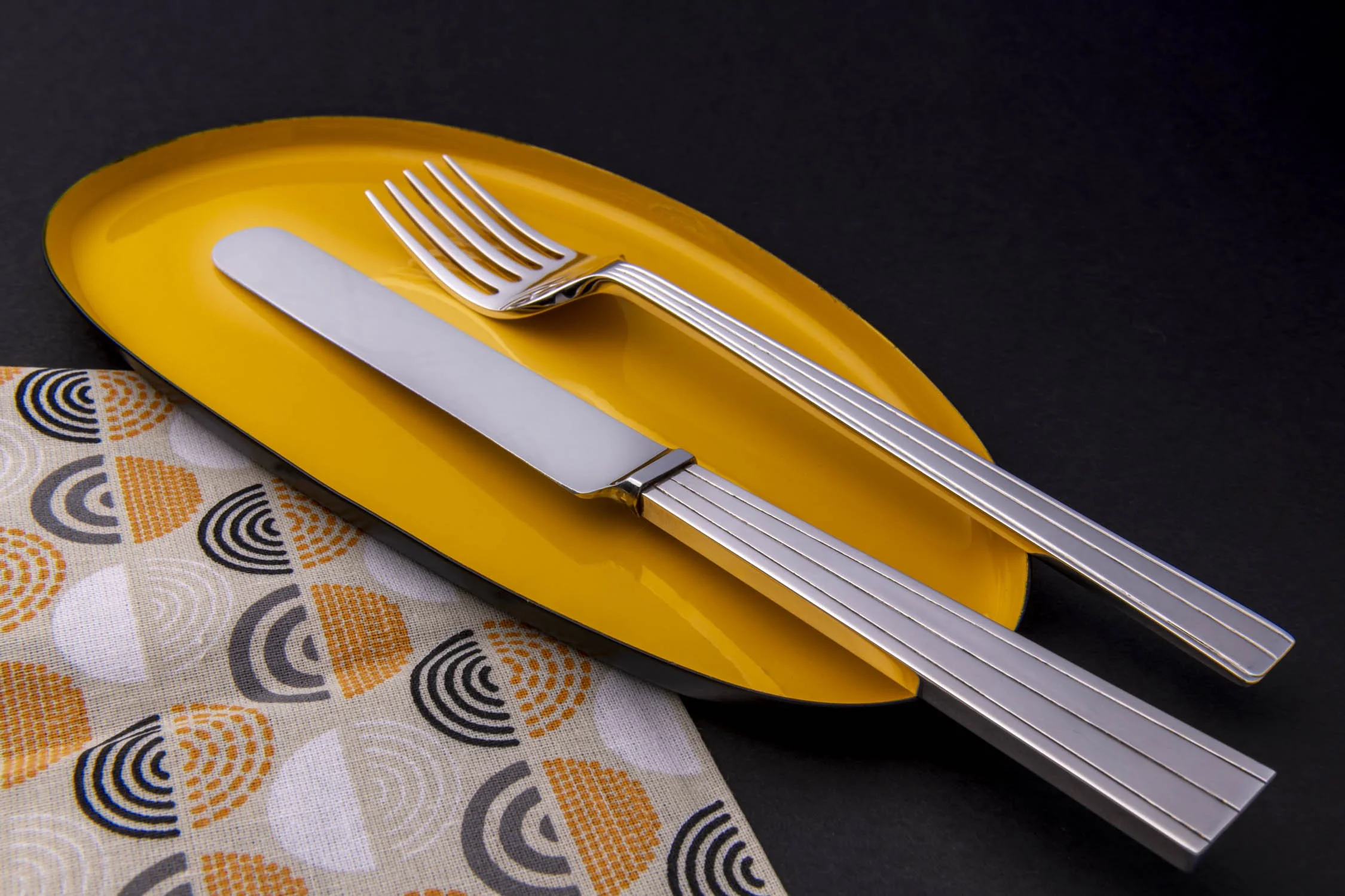 A yellow plate with a fork and knife on top, positioned on a cloth with a colourful pattern, on a black background.