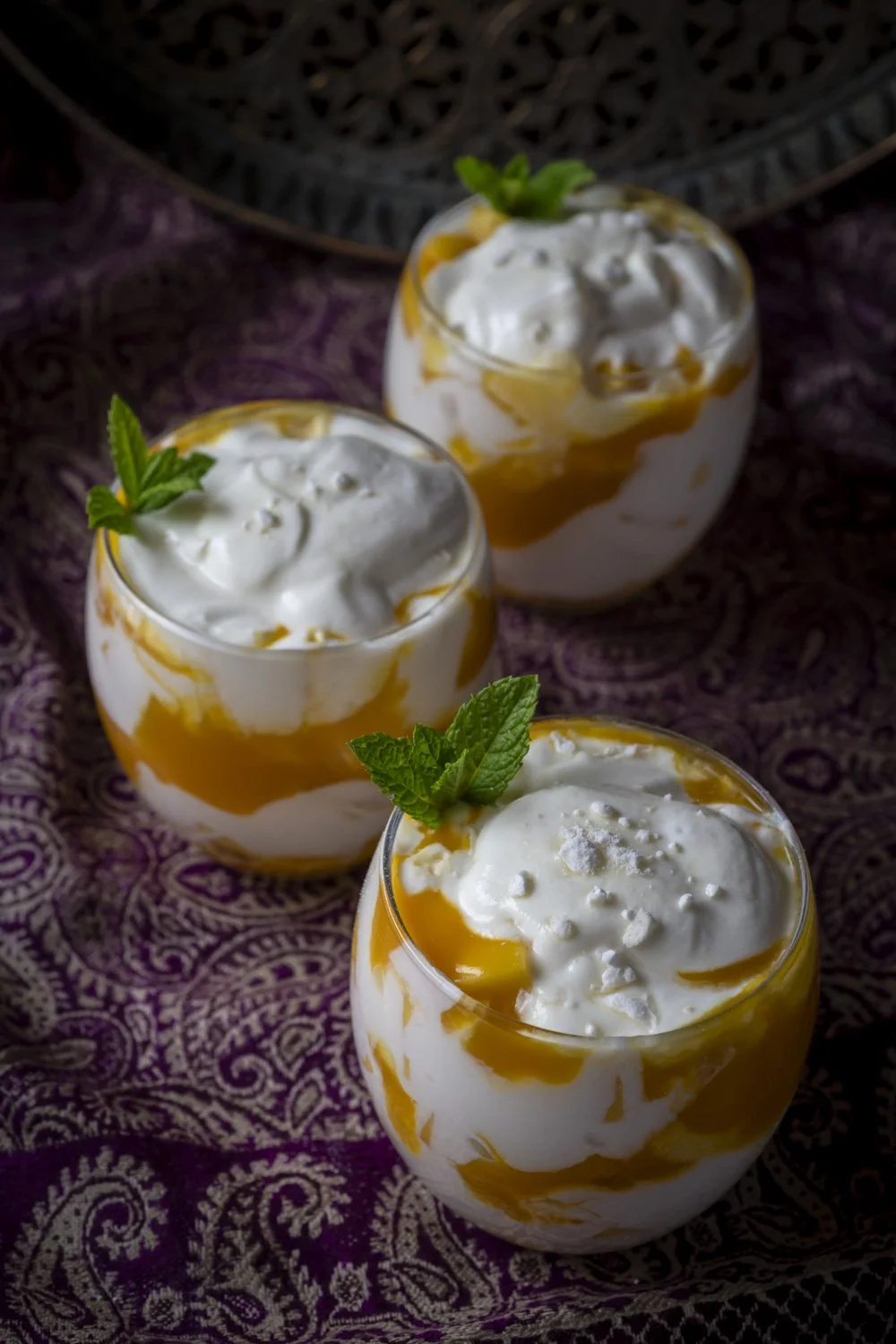 Three glasses of mango mousse topped with whipped cream and garnished with mint leaves, on a purple patterned cloth.