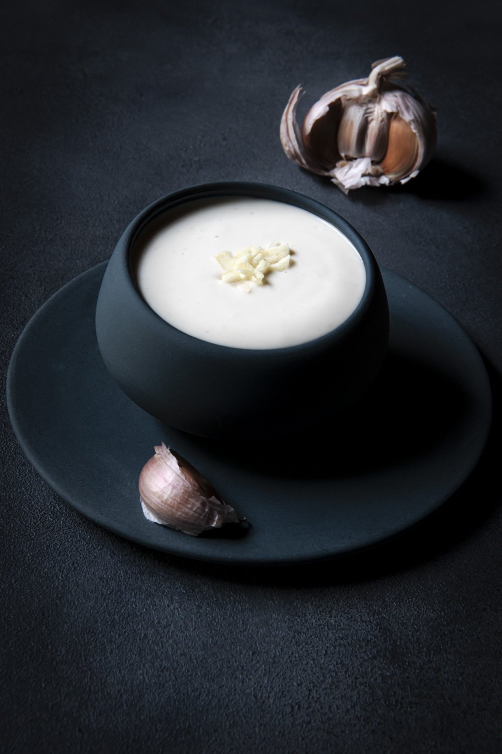 A bowl of creamy garlic sauce placed on a dark plate and table, with garlic cloves nearby.