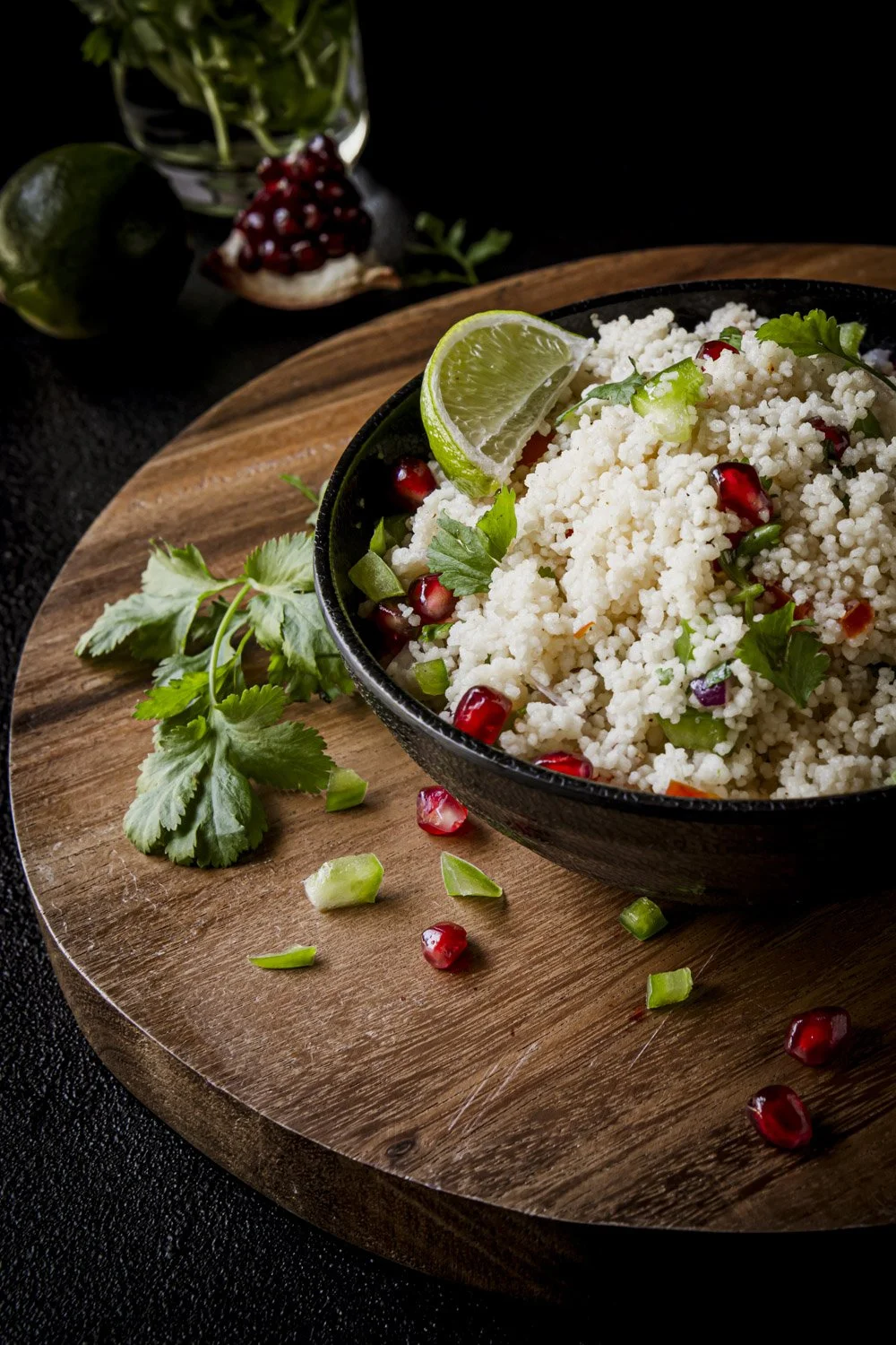 A bowl of couscous garnished with pomegranate seeds, coriander, chopped green onions, and a lime wedge, placed on a wooden serving board with additional herbs, pomegranate seeds, and chopped green onions scattered around.