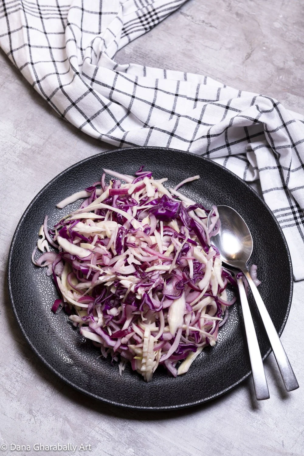 A black plate with shredded purple cabbage, placed on a light-coloured surface with a checkered tea towel nearby and a silver spoon and fork beside the cabbage.