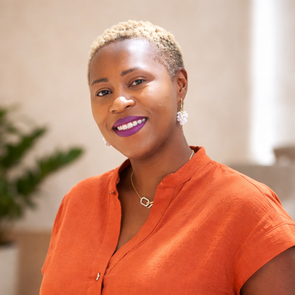 Portrait of a woman with short, bleached blonde hair, wearing purple lipstick, gold earrings, and a necklace with a circular pendant, dressed in an orange top, smiling indoors.
