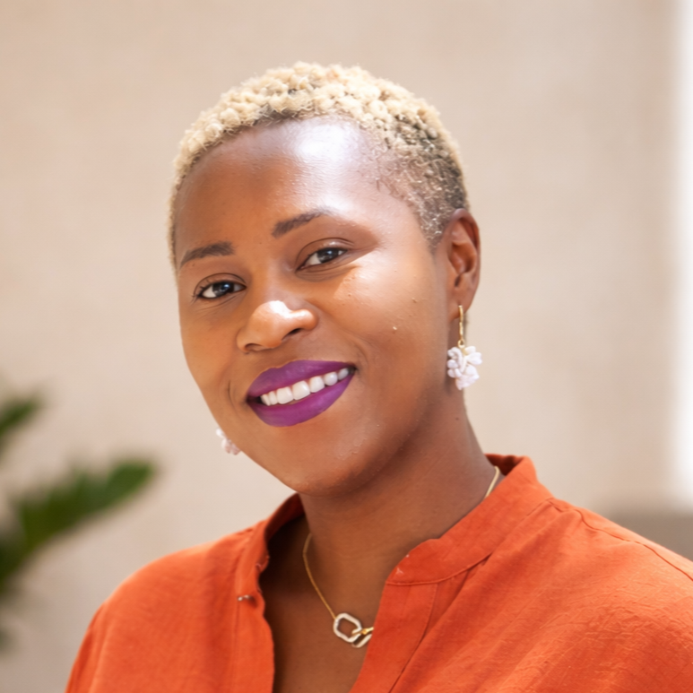 Portrait of a woman with short, bleached blonde hair, wearing purple lipstick, gold earrings, and a necklace with a circular pendant, dressed in an orange top, smiling indoors.