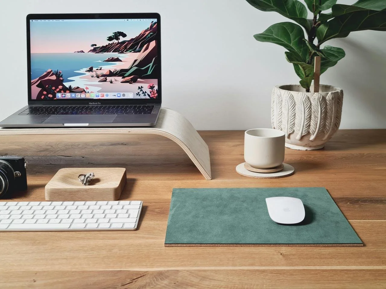 A tidy wooden desk with a MacBook Pro on a curved wooden stand, a wireless keyboard, and a mouse on a teal mouse pad. Next to the MacBook is a potted plant with large green leaves in a decorative white pot, a white ceramic mug on a coaster, and a small camera. A pair of glasses is on a wooden holder in front of the mouse.