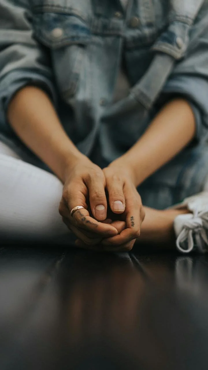 Person wearing a denim jacket, sitting with hands clasped, resting on black surface.