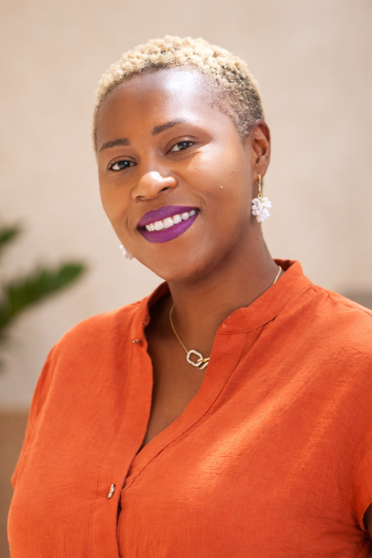 A smiling woman with short, blonde hair and purple lipstick, wearing gold earrings and a necklace, dressed in an orange blouse.
